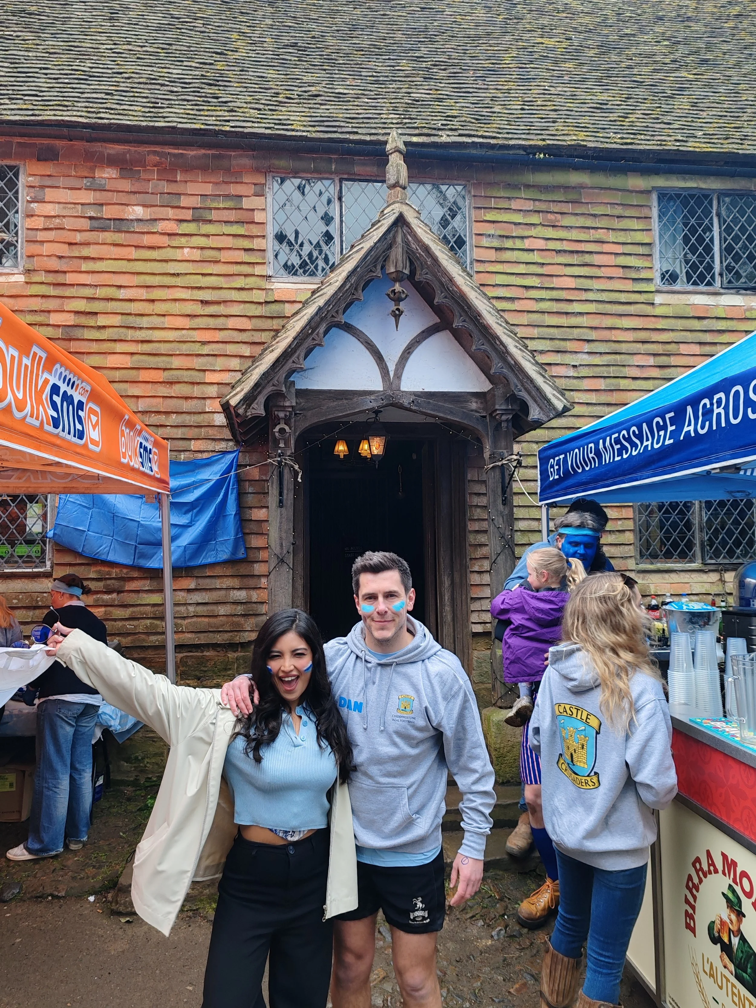Two people are smiling and posing for the camera in front of an old building with a wooden entrance and leaded glass windows. People are gathered around, some wearing colorful clothing, and some with painted faces, indicating a festive outdoor event.