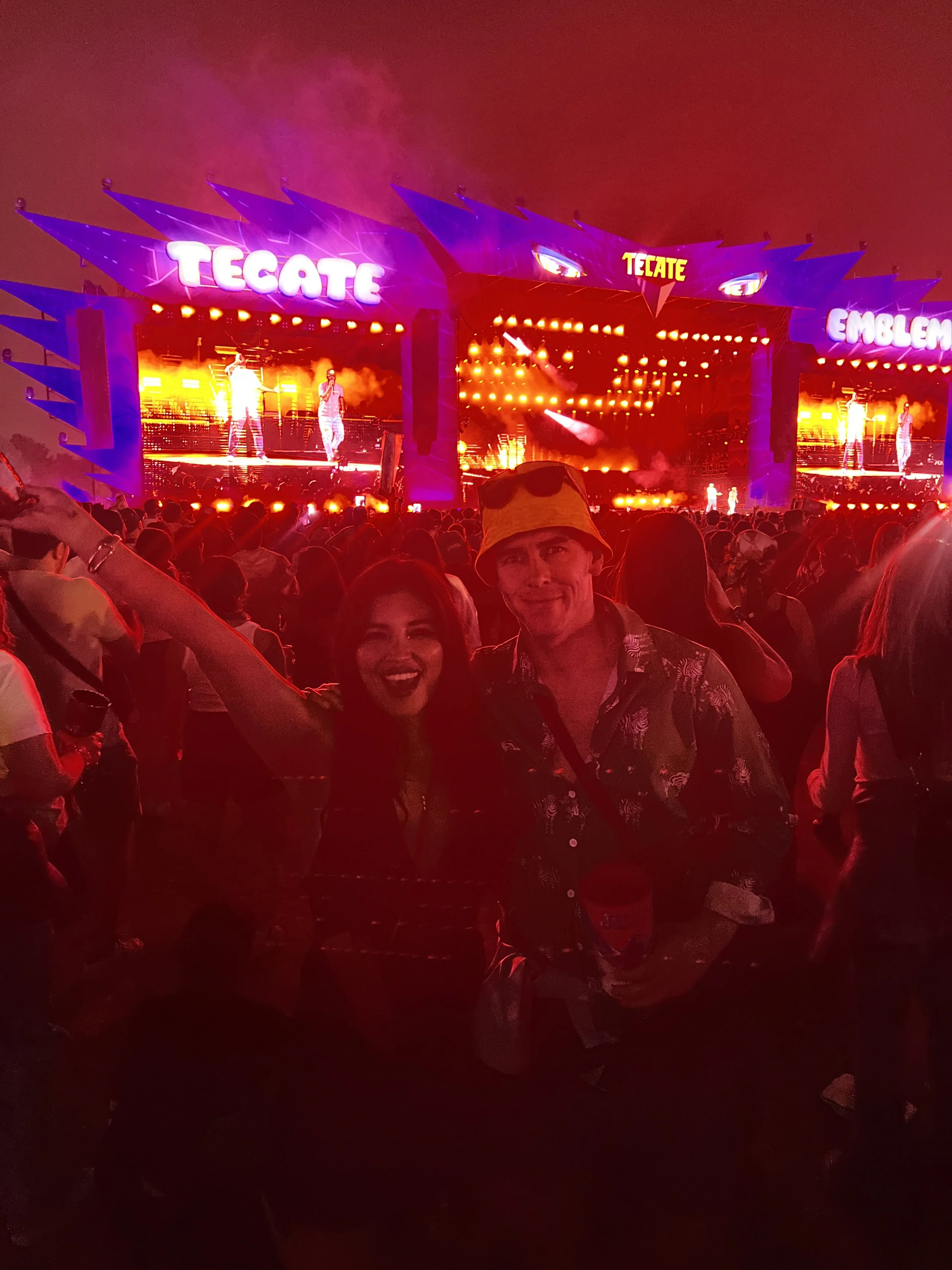 A concert stage with bright orange lights and a large screen displaying performers. The stage has prominent signs saying 'TEGATE' and 'EMBLEM'. The audience is gathered in front, with some people visible in the foreground.