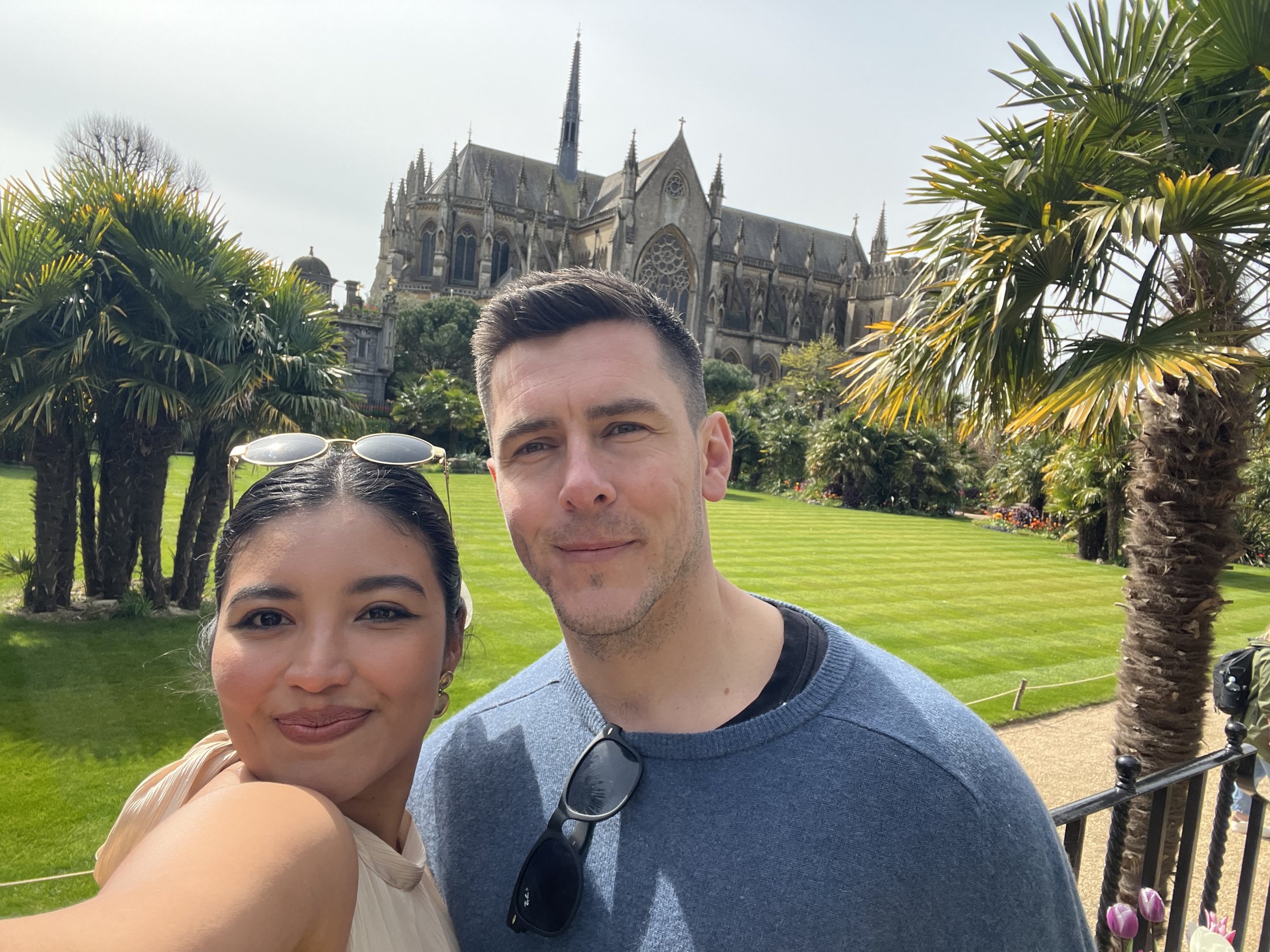 A woman and a man taking a selfie in a park with a Gothic-style cathedral in the background. The woman has dark hair, sunglasses on her head, and a light-colored dress. The man has short dark hair, glasses hanging from his shirt, and is wearing a blue sweater. There are lush green trees, grass, and blooming flowers around them.