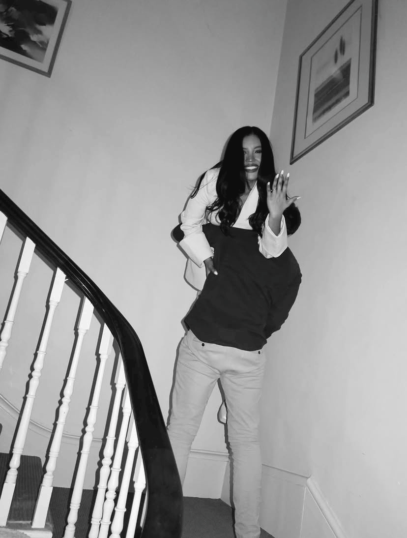 A woman with long dark hair is smiling and waving at the camera while standing on a staircase. She is wearing a light-colored blazer over a dark hoodie and light-colored pants.