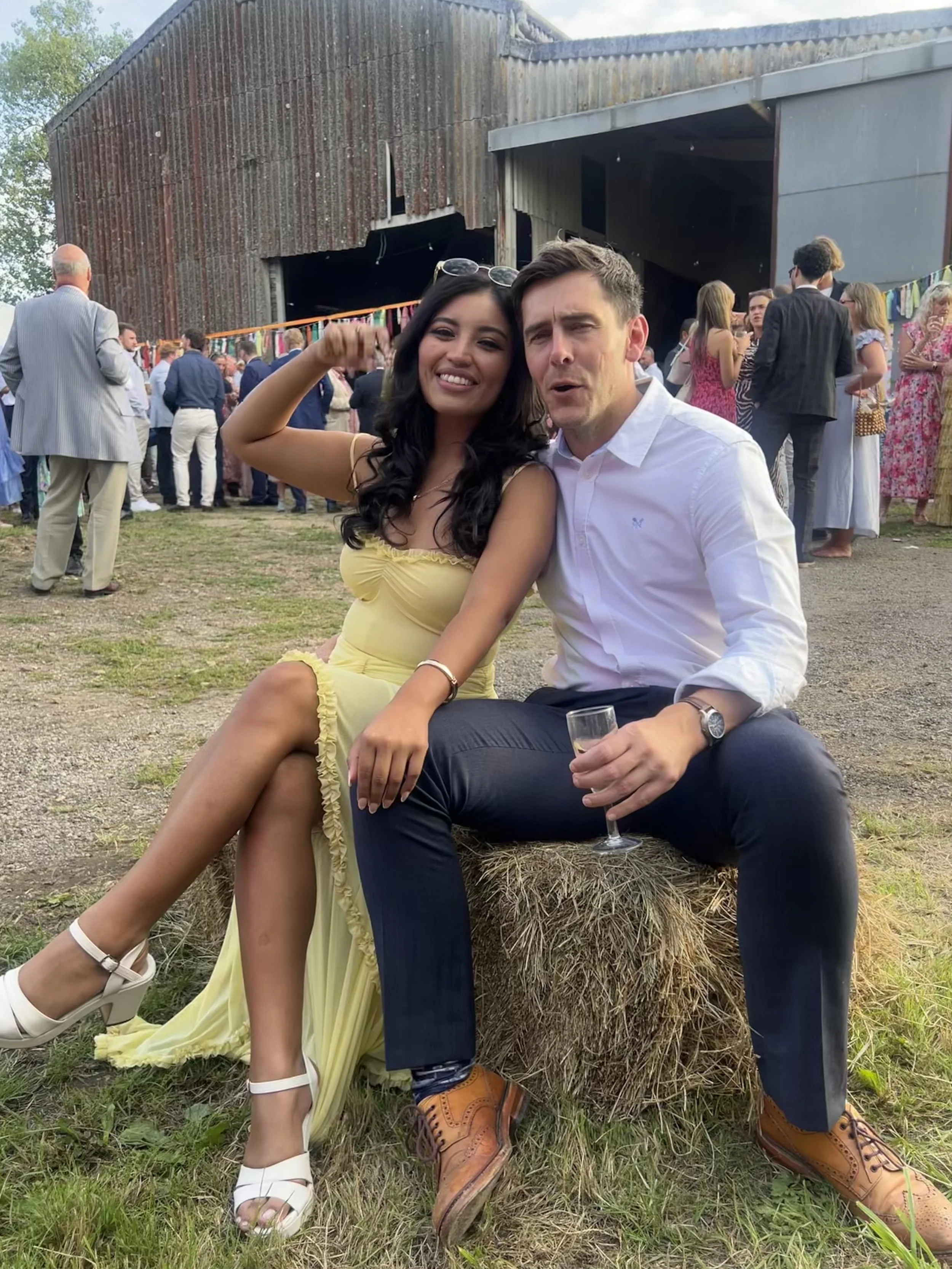 A woman in a yellow dress and a man in a white shirt are sitting outside on a hay bale at a social event. The woman is smiling and flexing her arm, while the man is holding a glass and has a surprised expression. There are many people in the background mingling and talking, with a rustic barn and colorful decorations behind them.