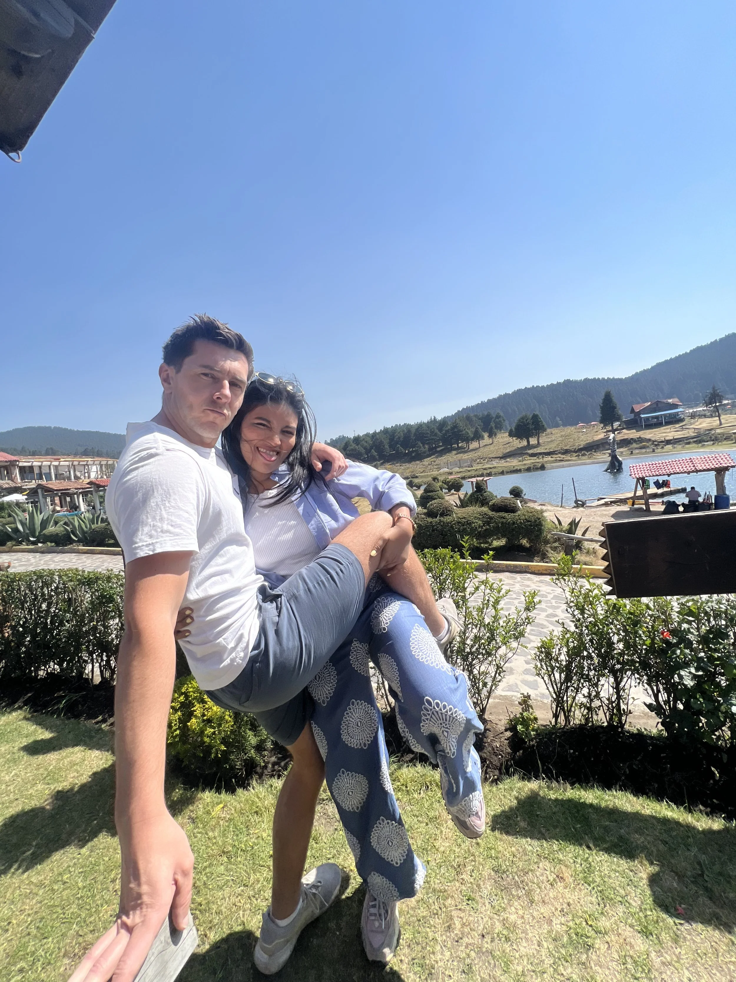 Two people, a man and a woman, sit on a grassy area outdoors, with a body of water and hills in the background. The man, wearing a white t-shirt and gray shorts, sits with a serious expression, while the woman, wearing a white shirt and patterned pants, smiles and leans into him. The sunny day and bright blue sky are visible, with some buildings and trees near the water.