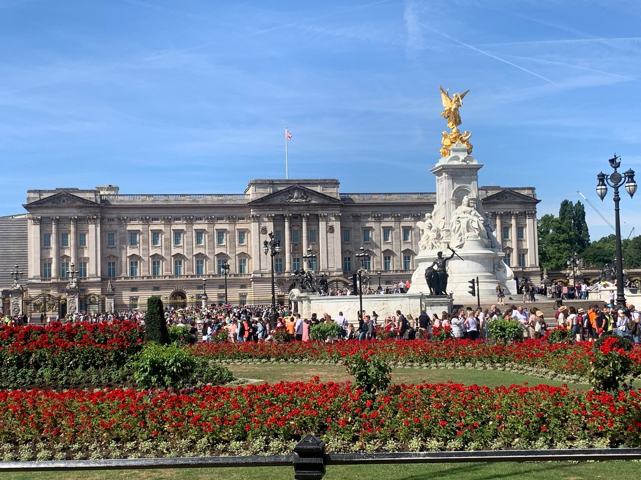 Crowd of people in front of a historic building with ornate architecture and a large white and gold statue on a tall pedestal, with a flag flying on the roof and a clear blue sky overhead.