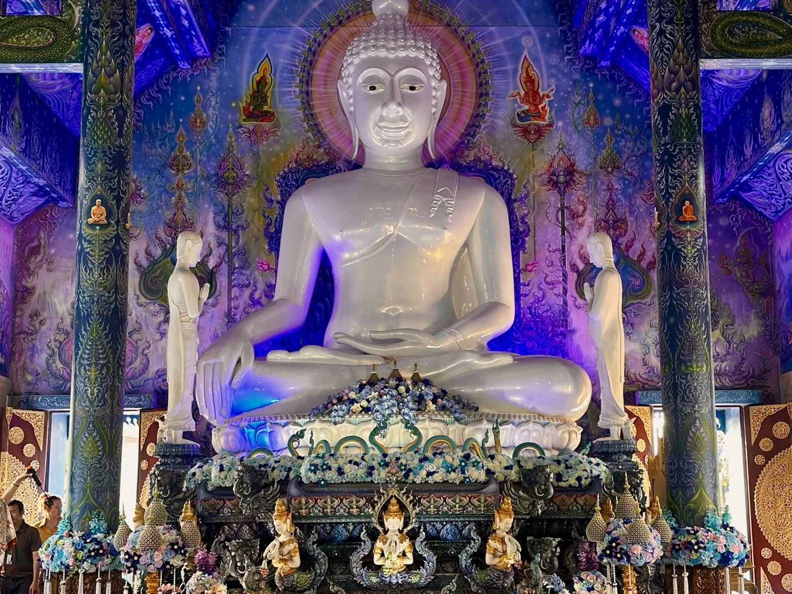 Large white Buddha statue in a decorated temple with colorful wall artwork, floral arrangements, and smaller statues.