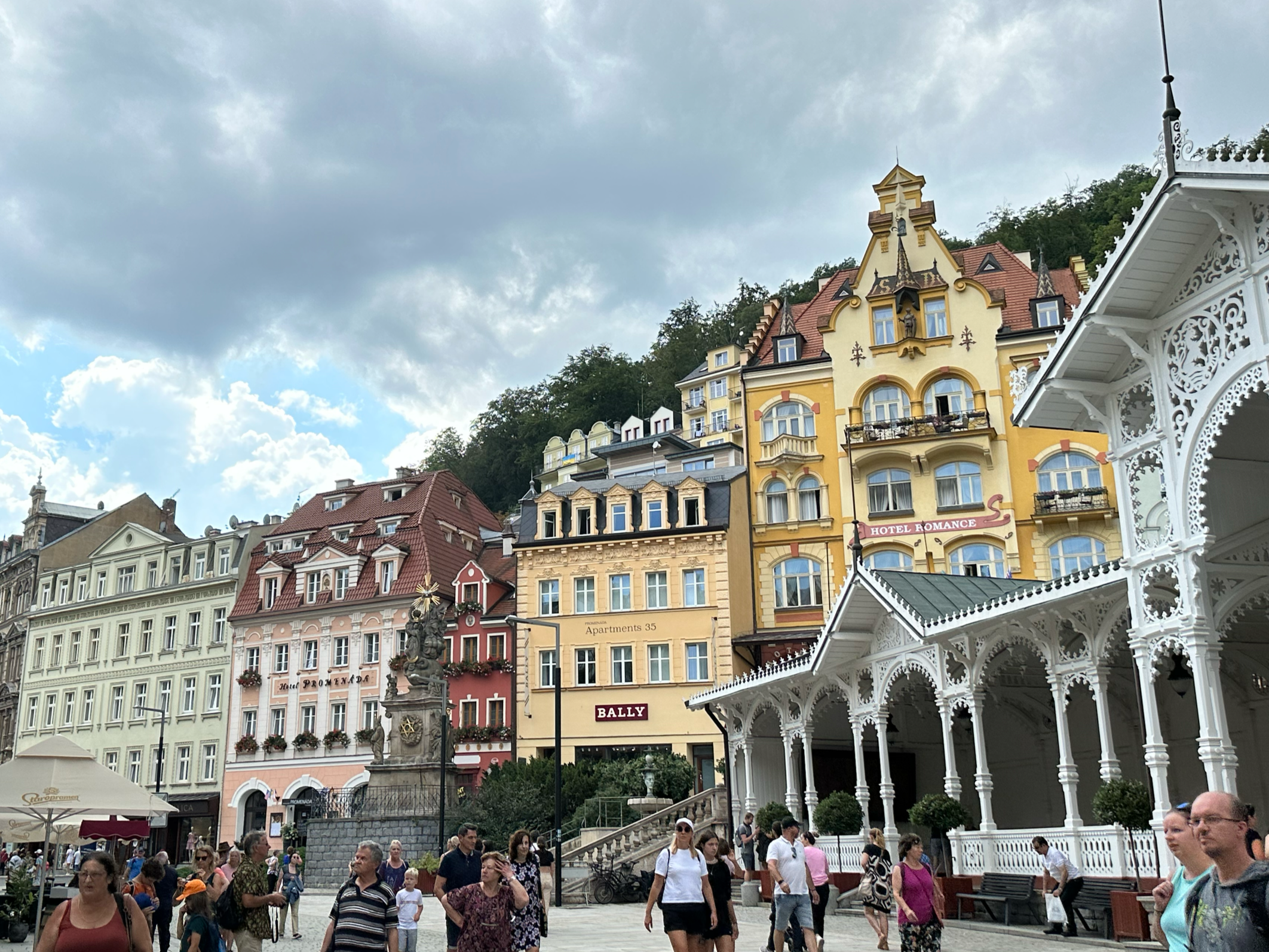 Crowd of people walking in a European town square with colorful historic buildings and a white ornate pavilion.