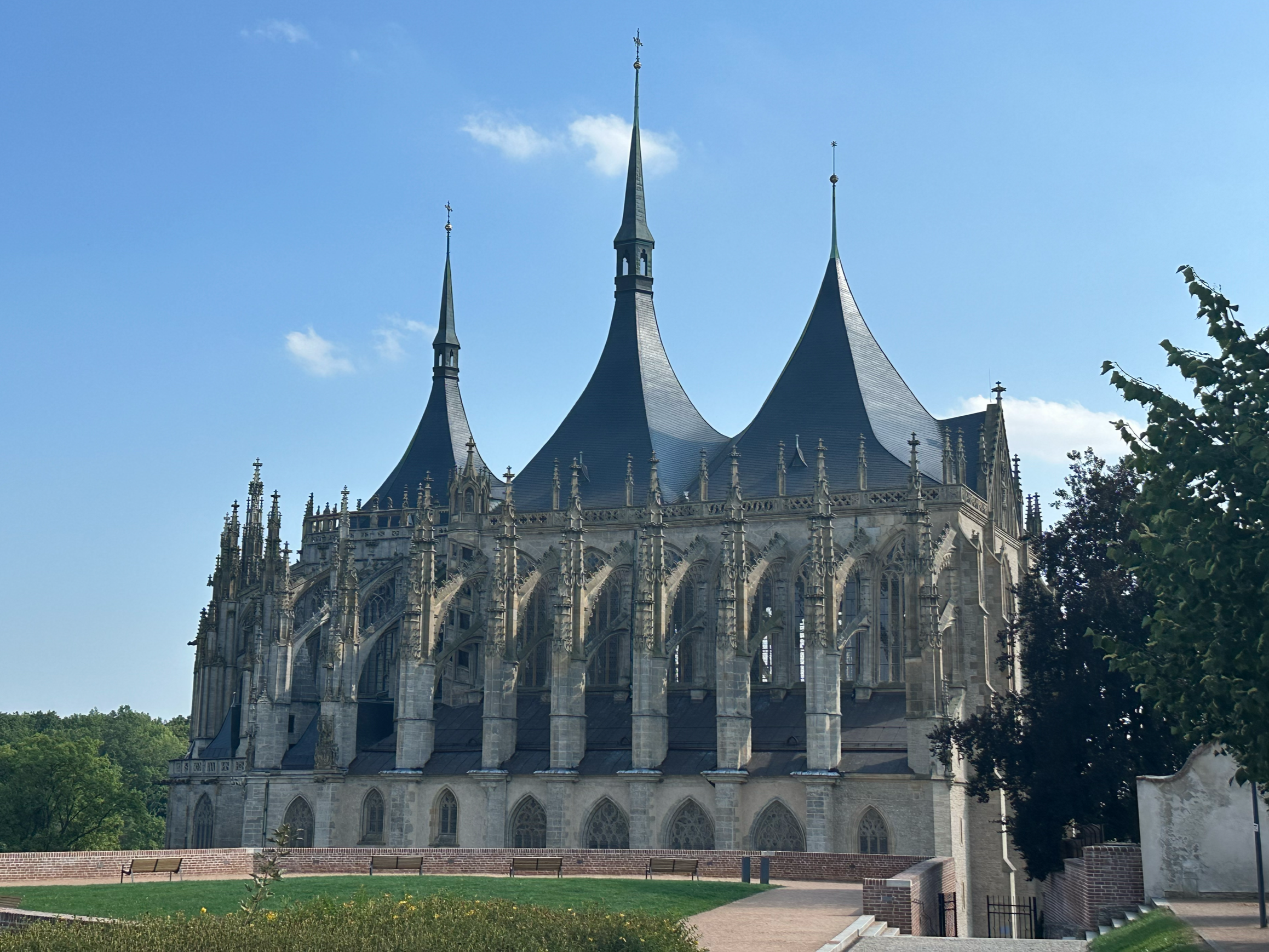 Front view of a historic Gothic-style cathedral with multiple pointed spires and detailed stone architecture, surrounded by a park with benches and trees, under a blue sky.