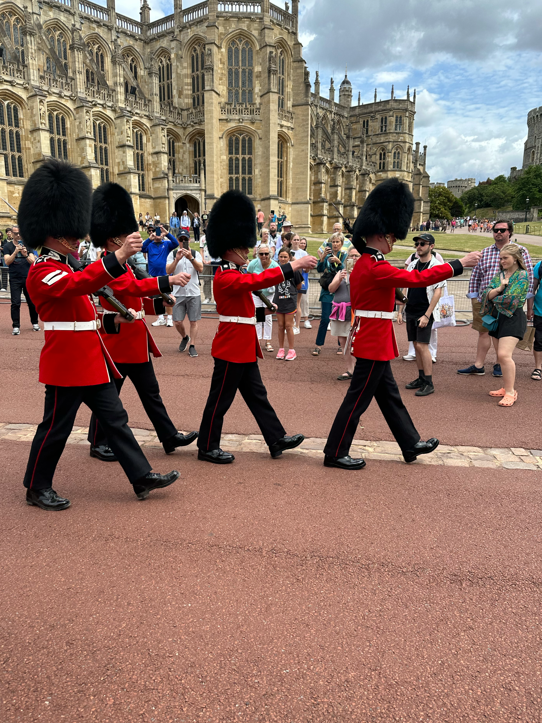 Four members of the Queen's Guard in red uniforms and tall black hats marching in front of Windsor Castle, with tourists watching and taking photos.