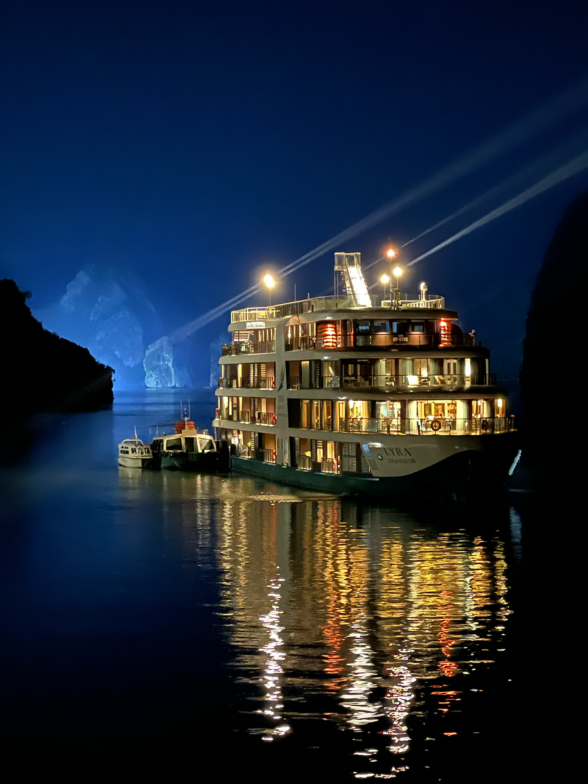 A nighttime scene of a boat docked in a harbor with illuminated buildings reflected in the water, and a mountain with clouds and a streak of lightning in the background.