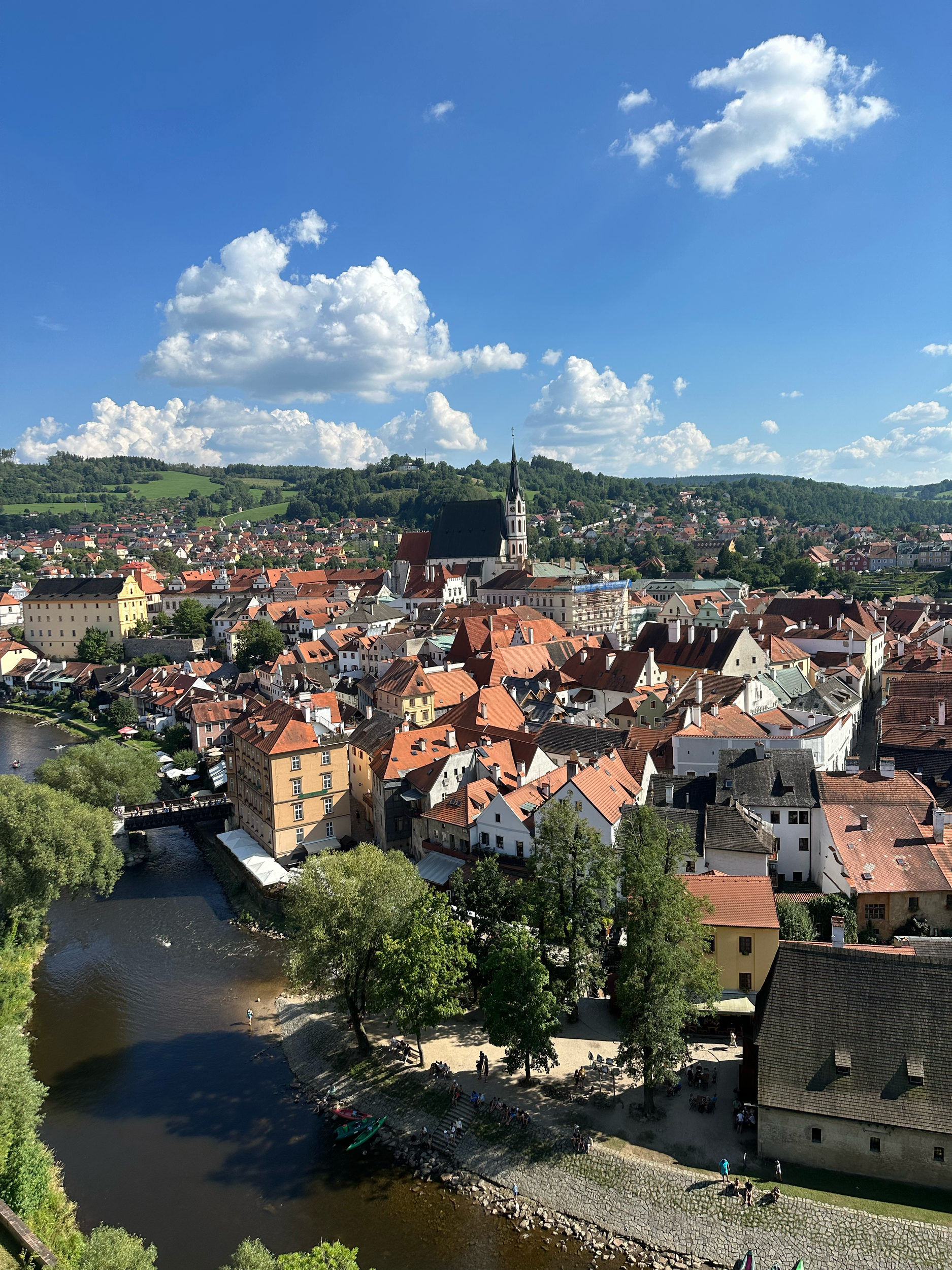 A scenic view of a European town with red-tiled roofs, church steeples, and a river running through the town, surrounded by green hills and blue sky with clouds.