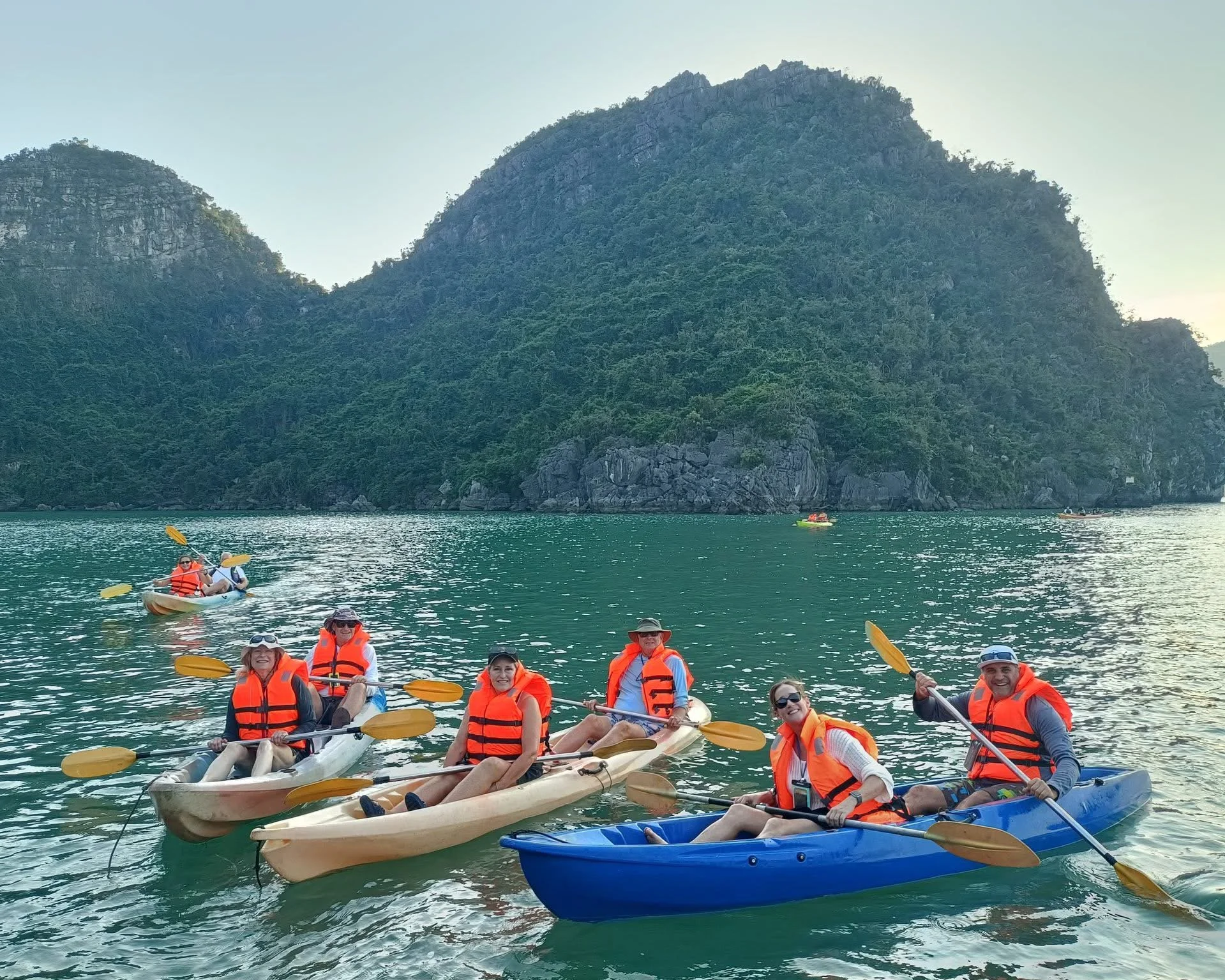 Group of people in kayaks on a lake with lush green mountains in the background, wearing orange life jackets and holding paddles.
