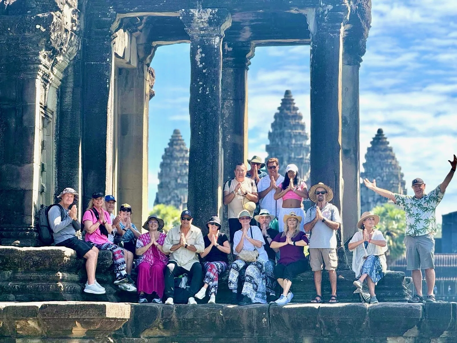 Group of tourists sitting and standing on ancient stone ruins, praying with hands pressed together, in front of temple towers at Angkor Wat in Cambodia, with blue sky and clouds in the background.