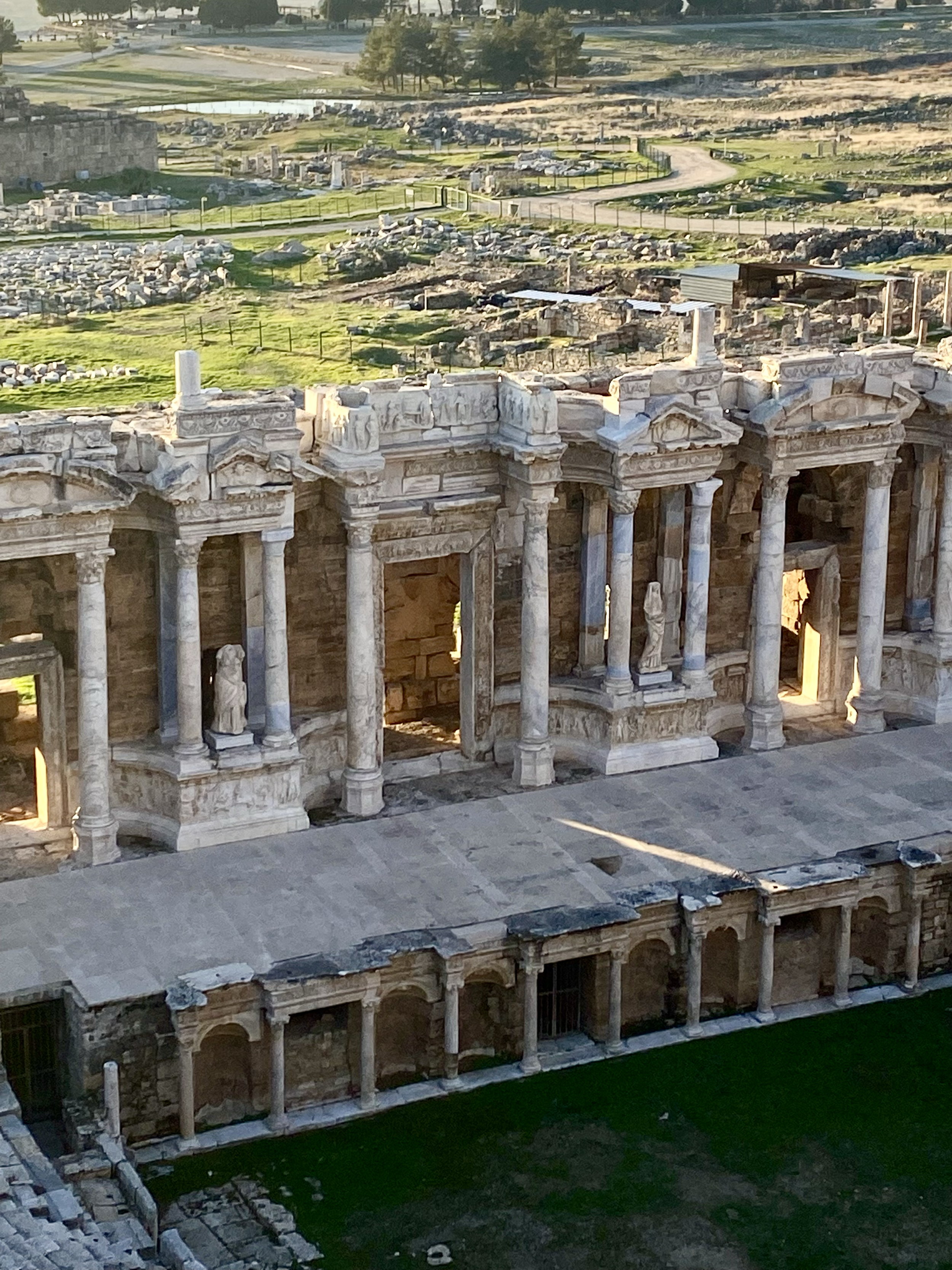 Ancient Roman theater ruins with tall marble columns and intricate carvings, set against a backdrop of green grass and distant trees.