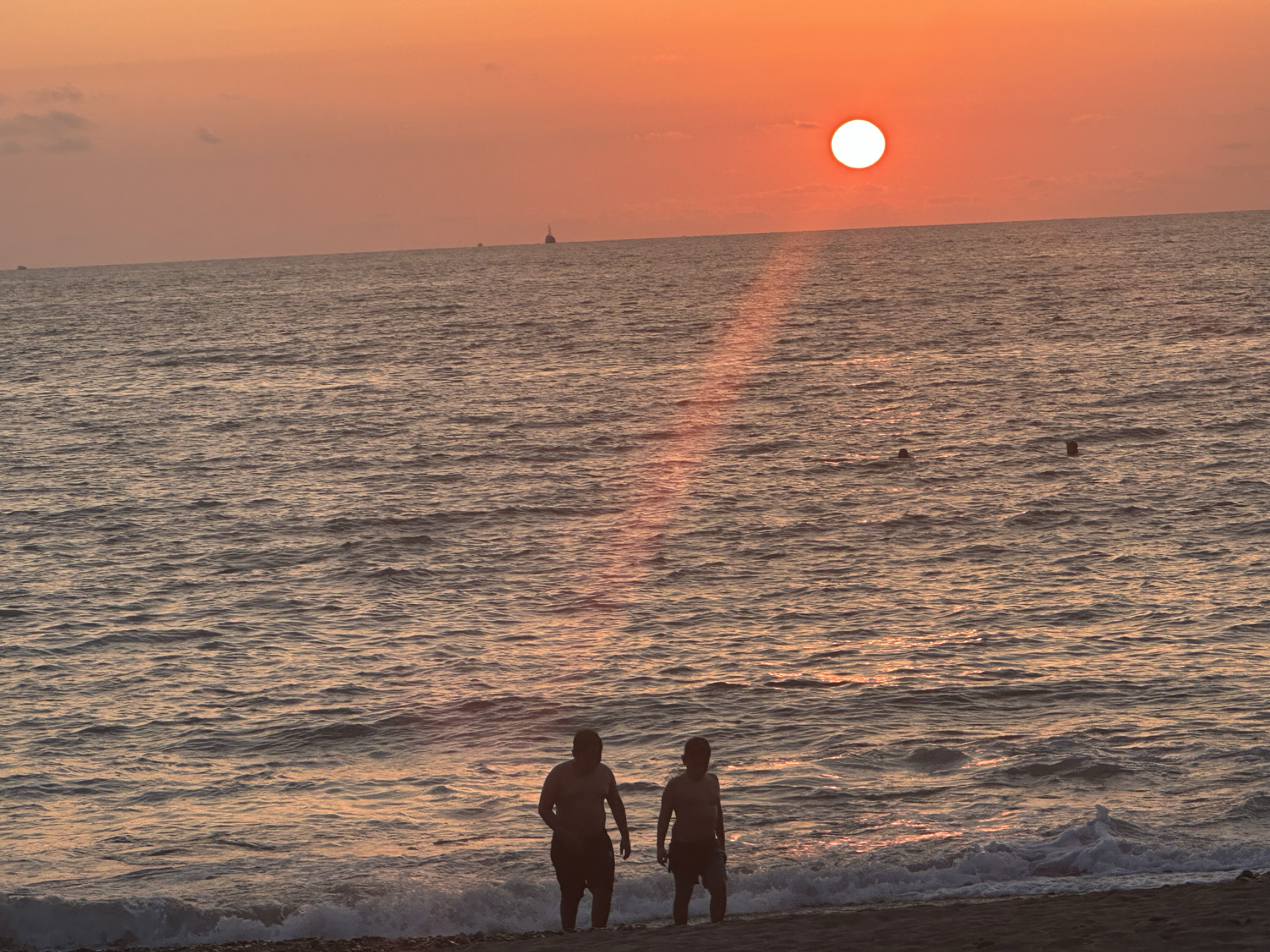 A sunset over the ocean with an orange and pink sky, the sun near the horizon, and two people walking along the shoreline.