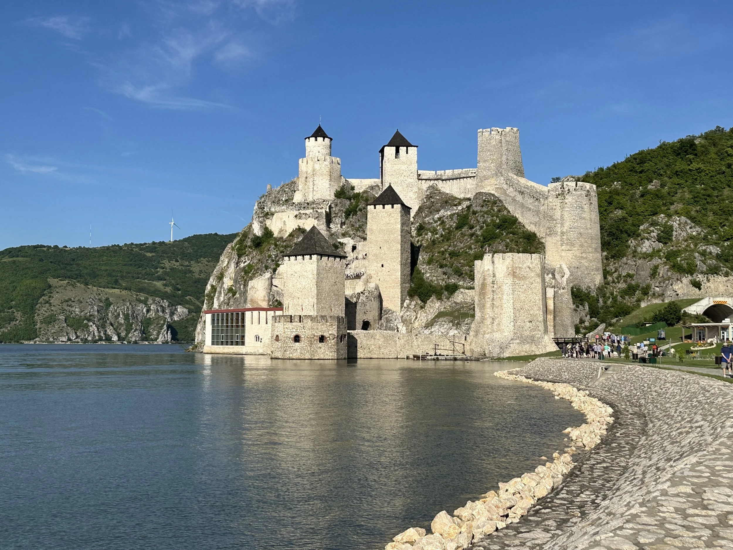 A scenic view of a historic stone castle on a hill, overlooking a body of water, with a walkway and group of people walking nearby. The sky is clear and blue, with wind turbines visible in the distance.