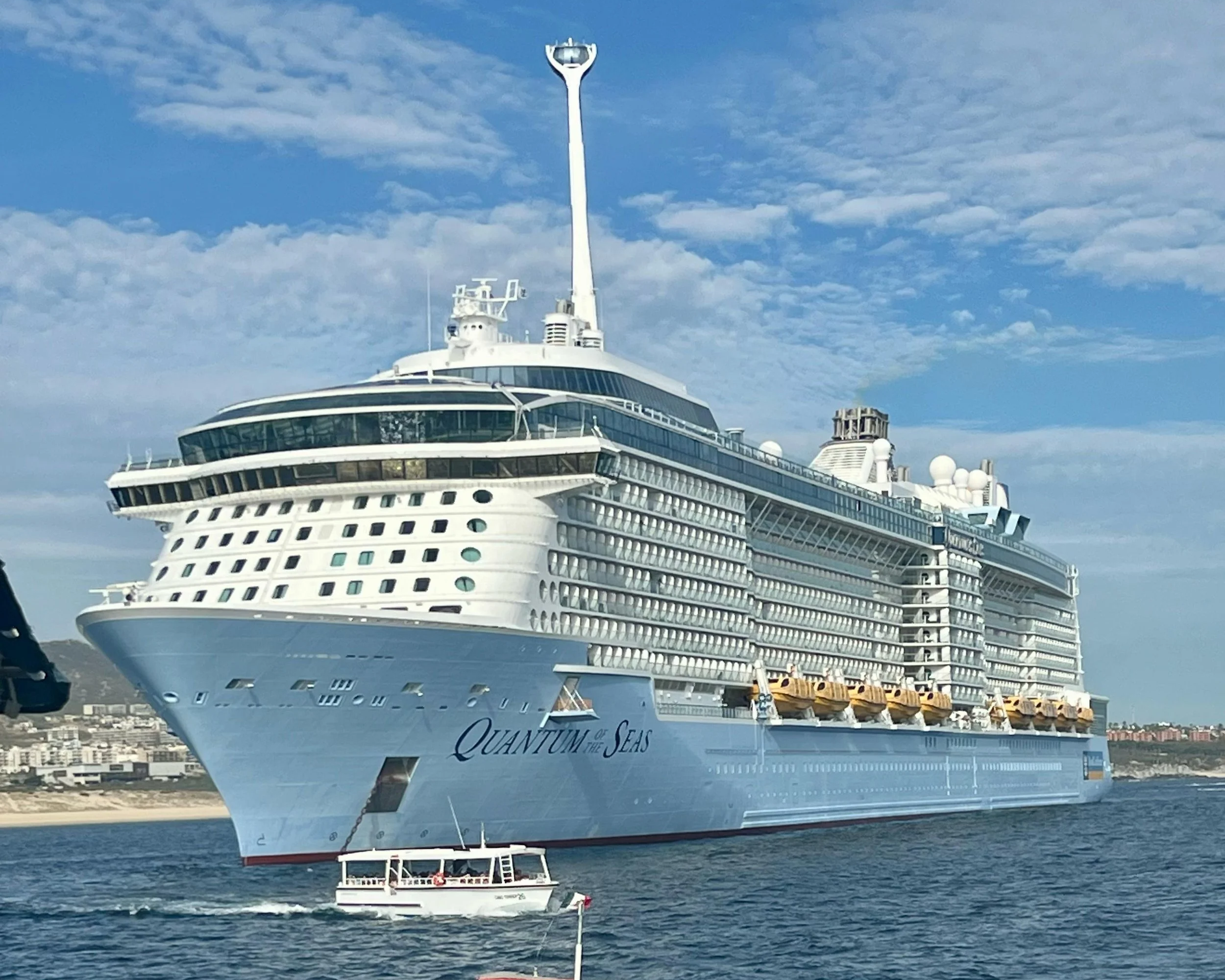 A large white cruise ship named Quantum of the Seas docked in a harbor with a cityscape in the background. The ship features multiple decks, a tall antenna, and various amenities on the upper decks, with a clear blue sky and some clouds overhead.