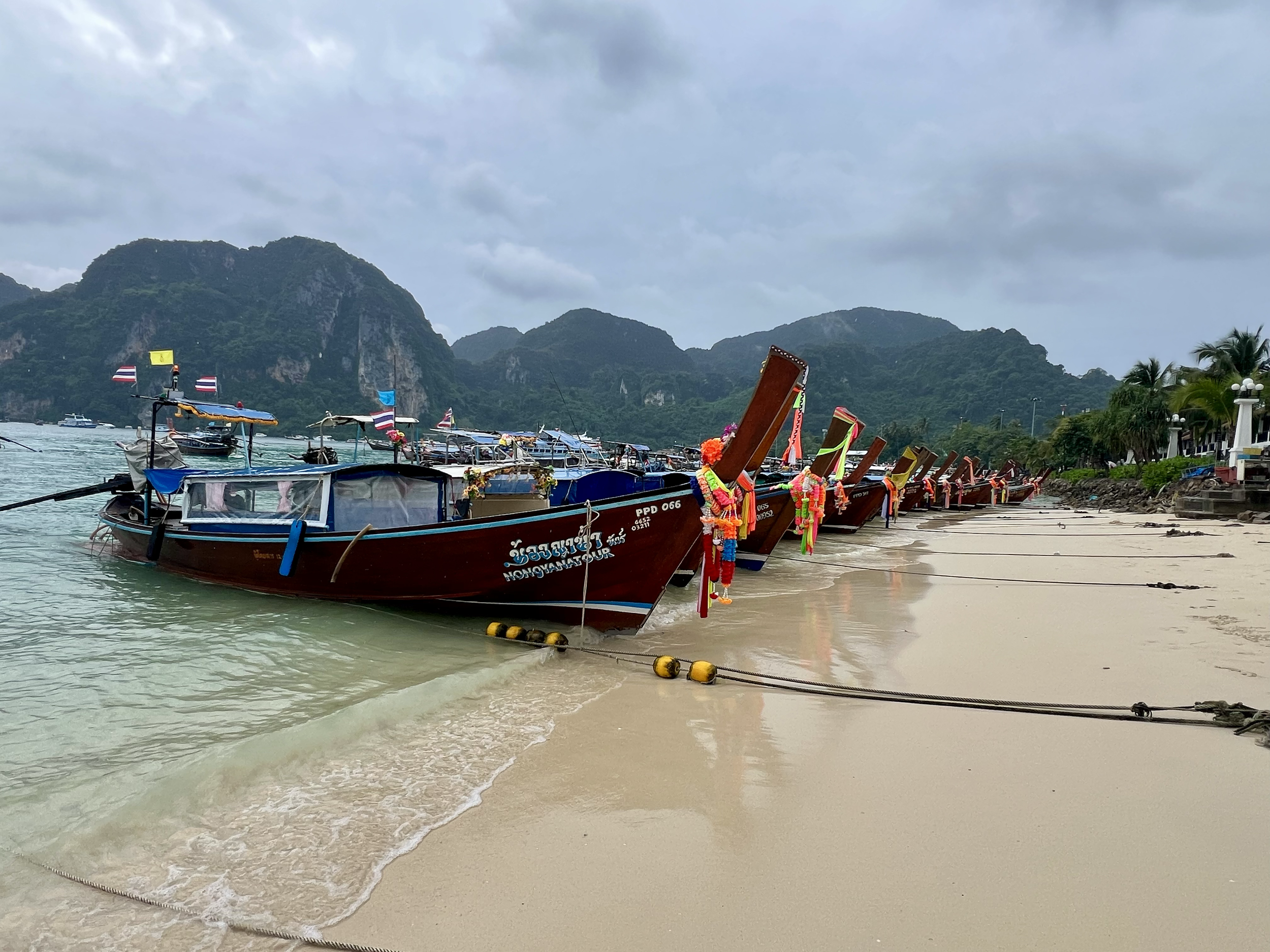 Beach with several traditional longtail boats docked along the sandy shore, mountains in the background under cloudy sky, and palm trees on the right.