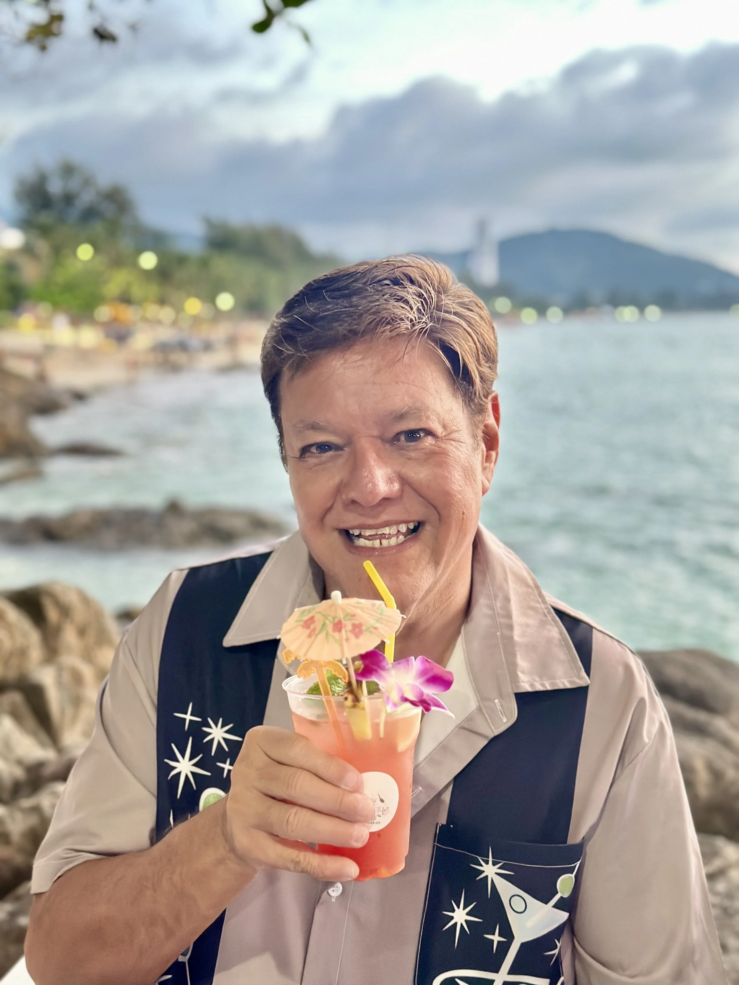 A smiling man holding a tropical cocktail with a small umbrella and orchid flower, at a seaside location with cloudy sky and mountain in the background.