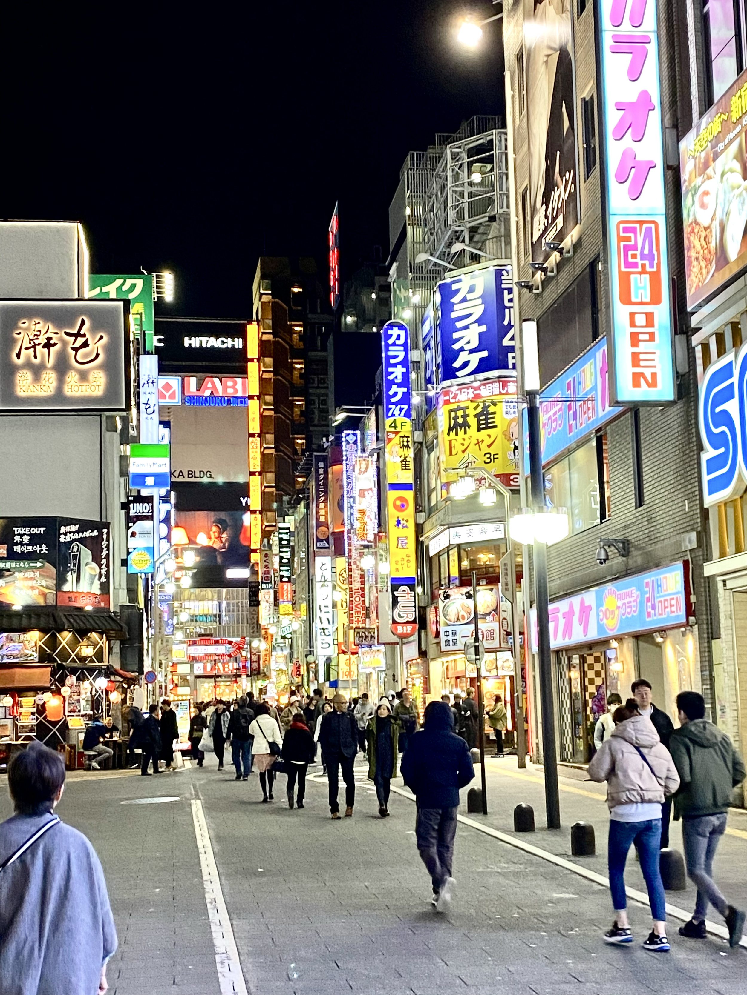 A busy city street at night with neon signs and many pedestrians walking along the sidewalks.