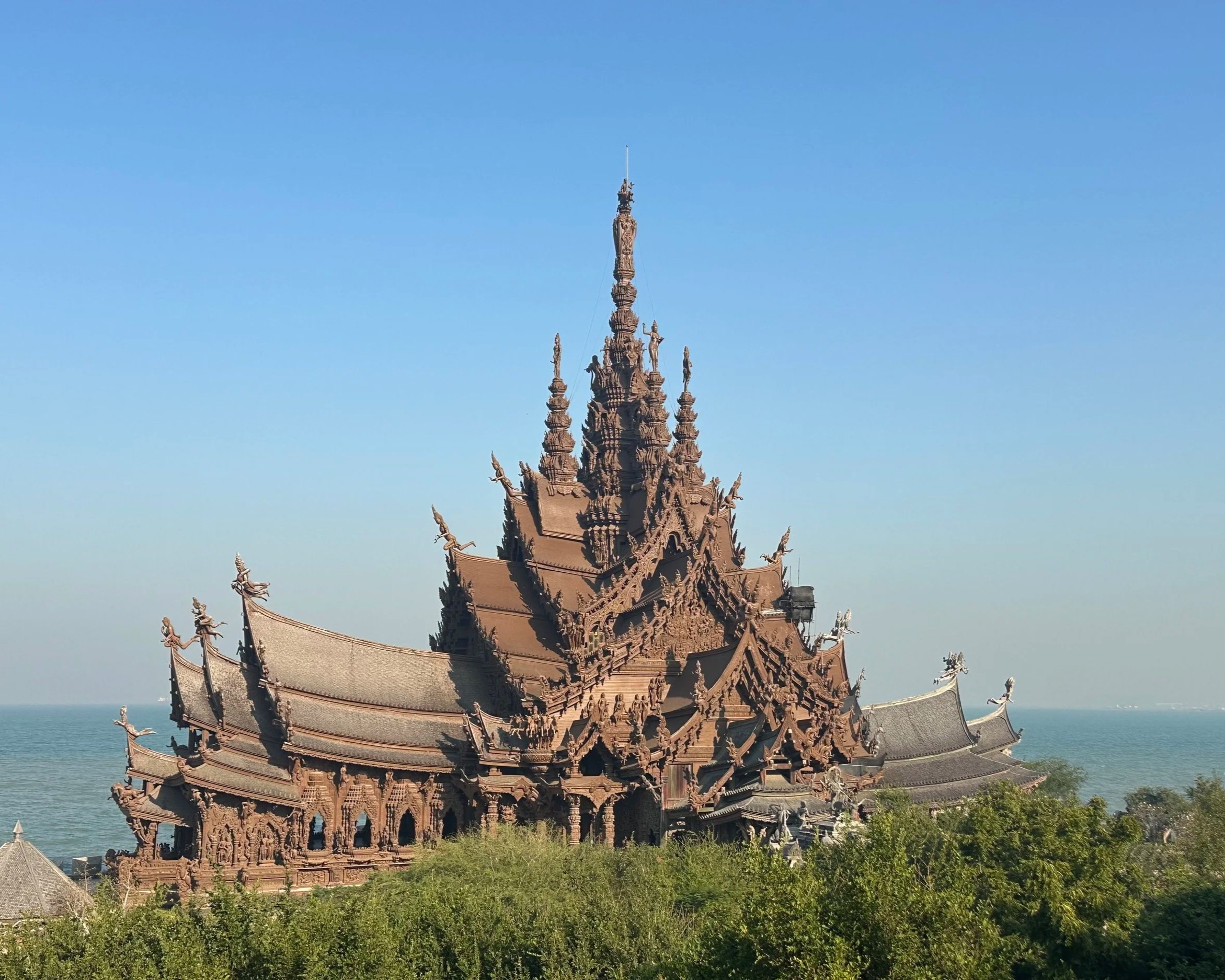 An ornate, traditional wooden temple with intricate carvings and sculptures, located by the water under a clear blue sky.