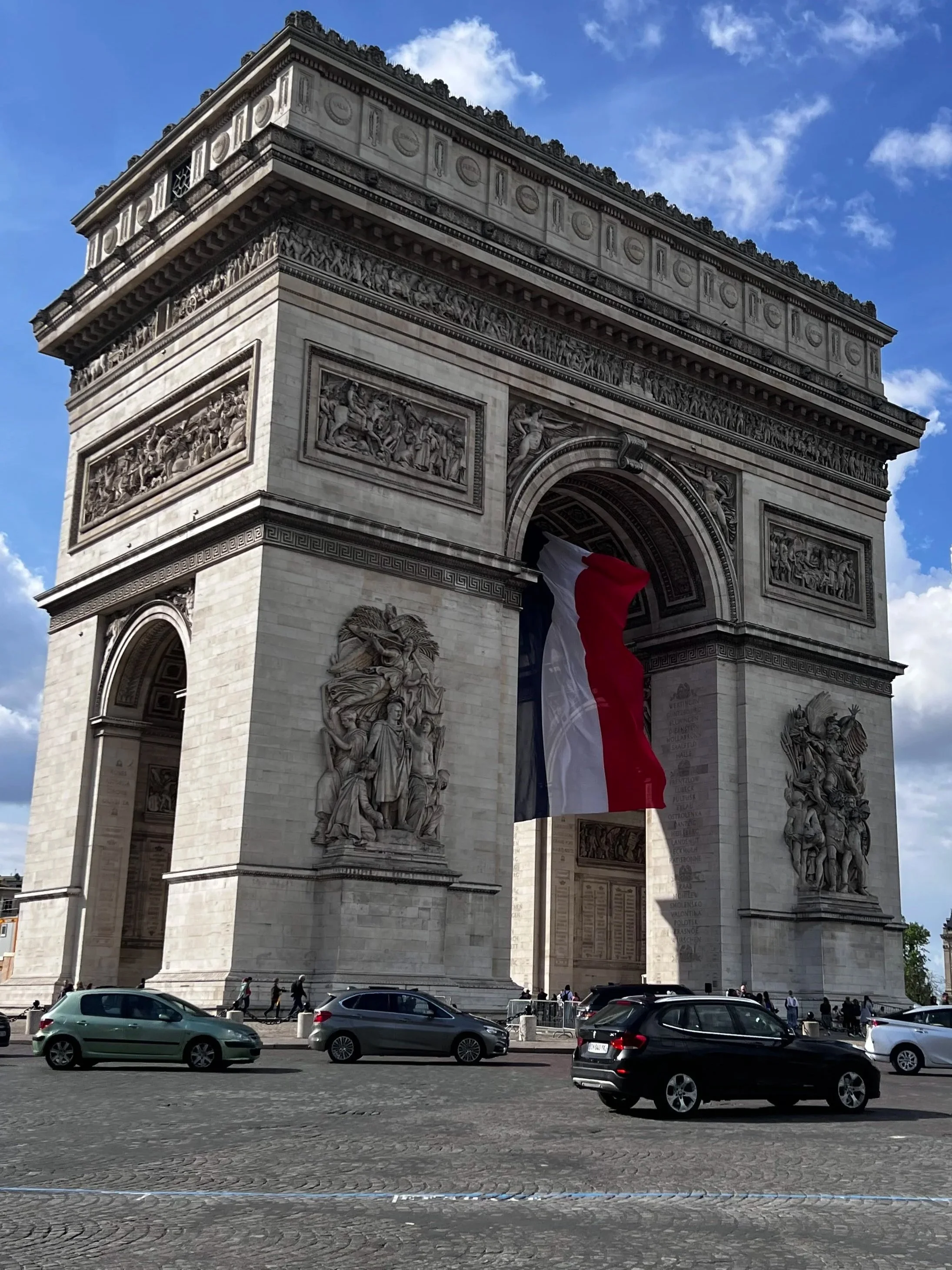 The Arc de Triomphe, a large Parisian monument, decorated with detailed sculptures and carved reliefs, and draped with the French flag. Vehicles and pedestrians are visible on the street in front of it, with a partly cloudy sky above.