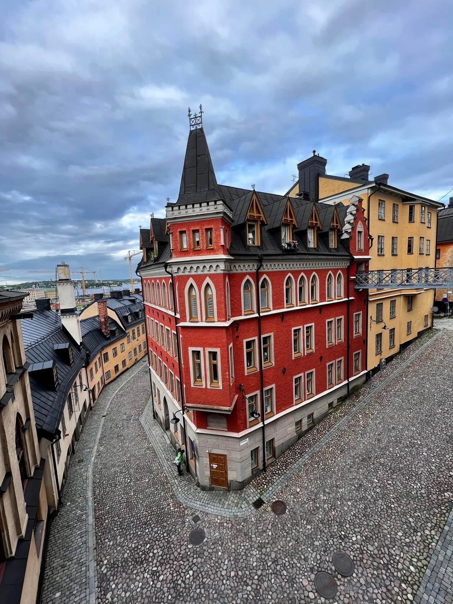 A colorful historic building with a pointed tower, ornate windows, and decorative trim, located on a cobblestone street under a cloudy sky.