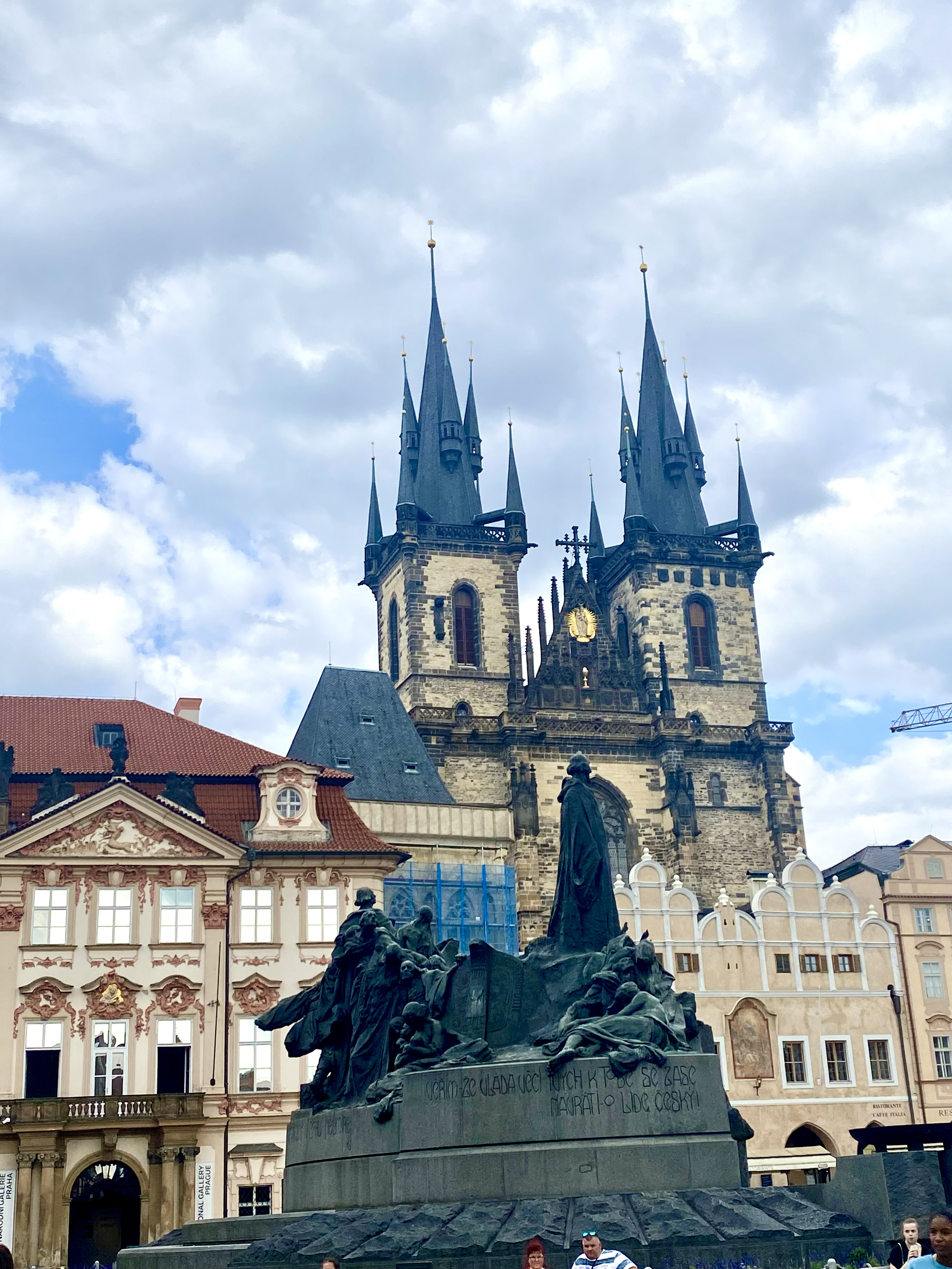 A church with tall spires, pointed arches, and Gothic architecture, set against a partly cloudy sky. In the foreground, a monument with statues and inscriptions, and people walking nearby.