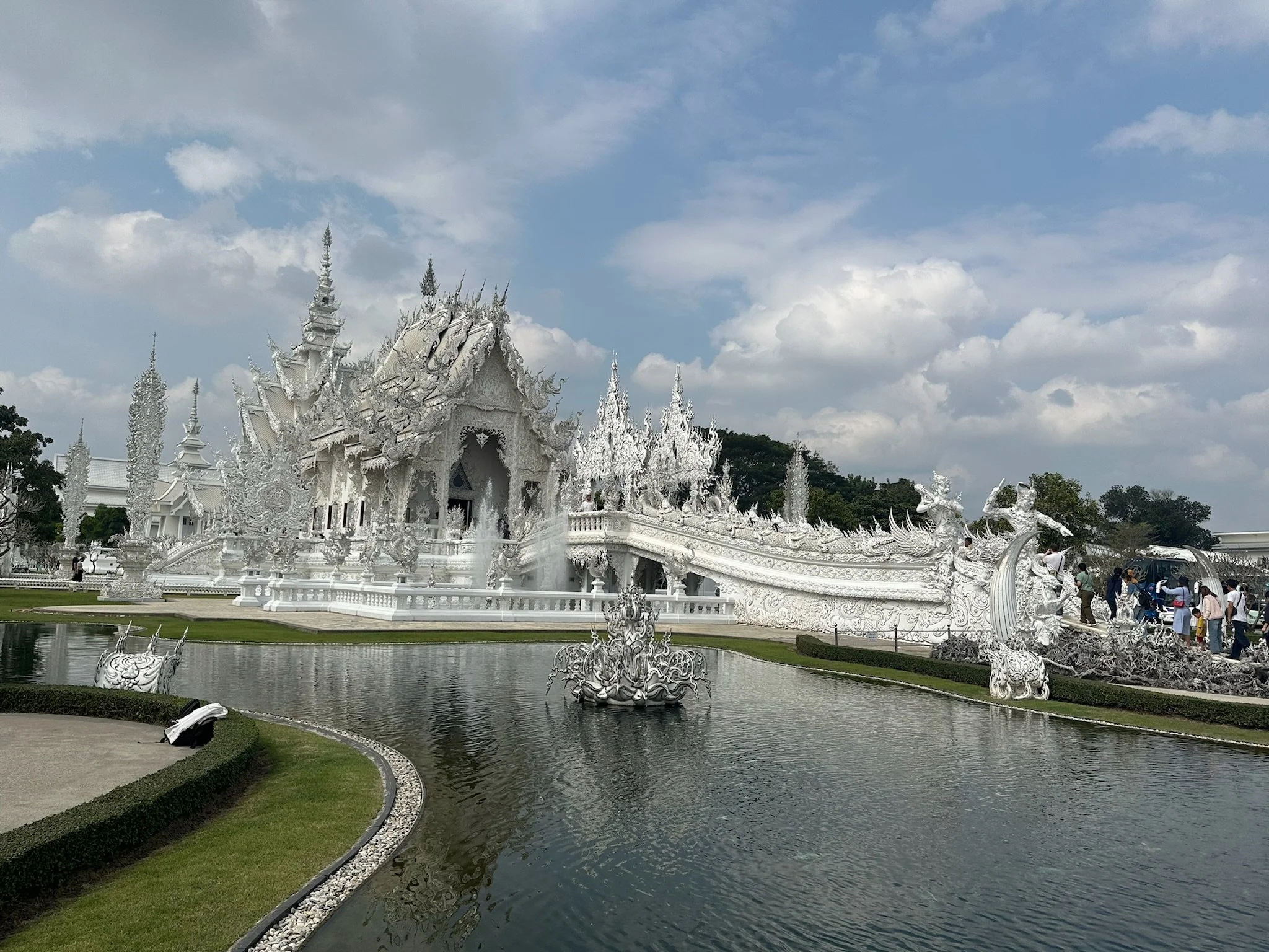 An ornate white temple with intricate carvings and pointed spires, surrounded by a pond with decorative dragon sculptures, under a partly cloudy sky. Visitors are walking around the temple grounds.