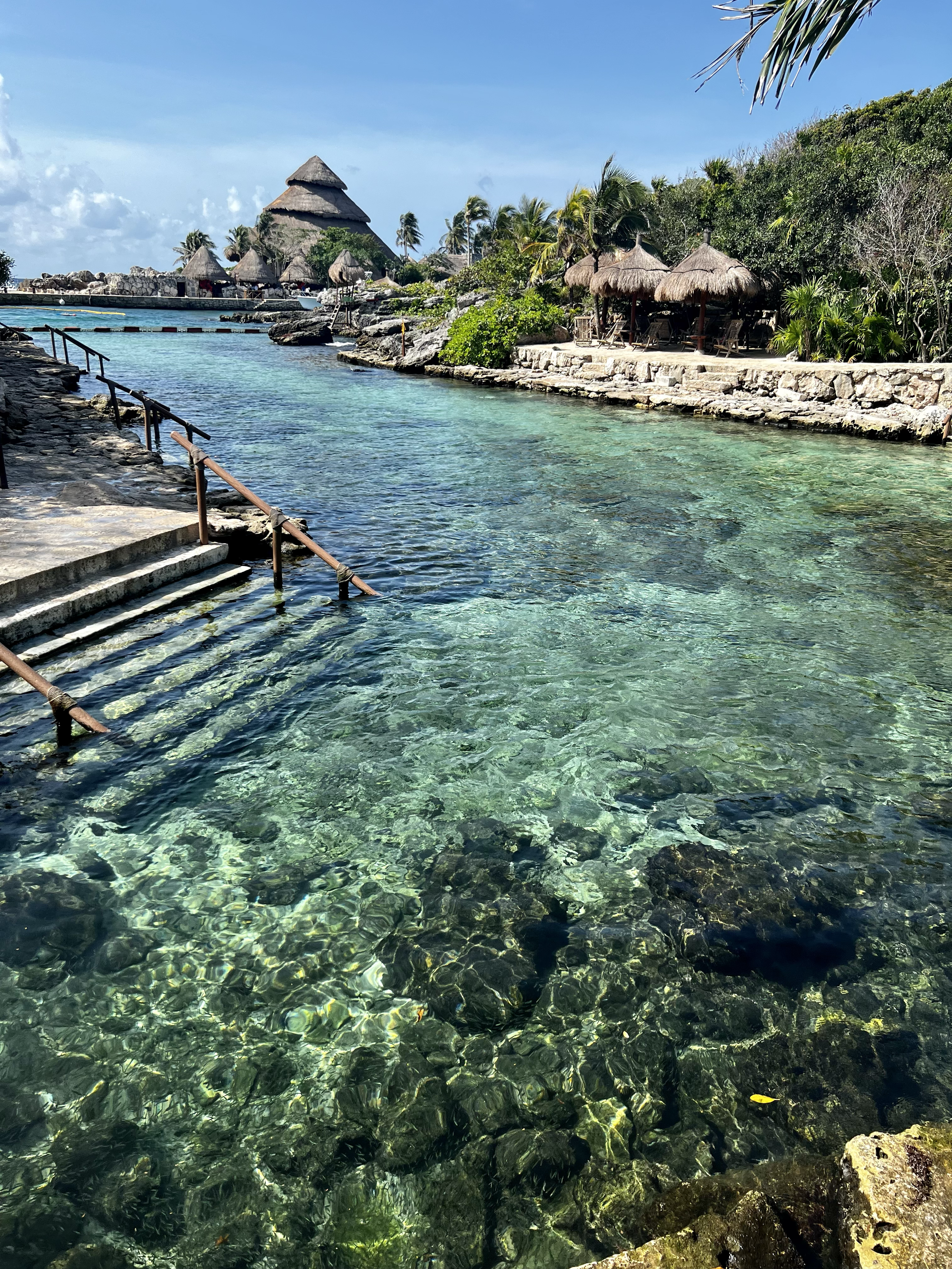 Tropical beach with clear water, thatched roof huts, lush green trees, and bright sunny sky.