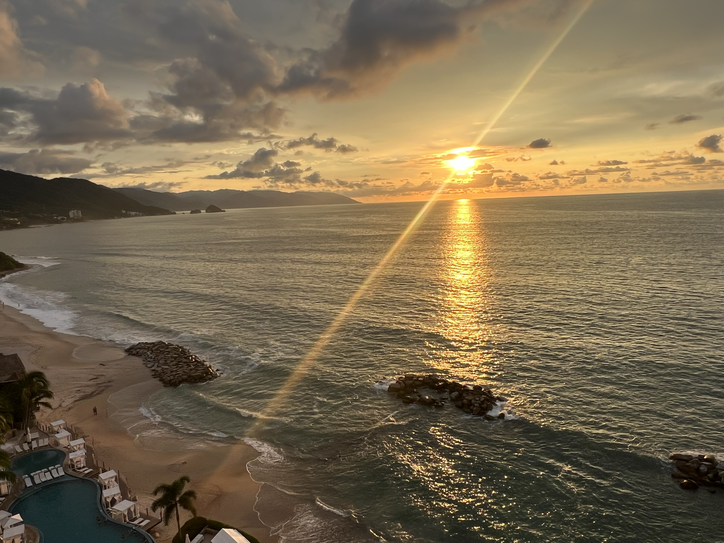 A sunset over the ocean with partly cloudy skies, a distant shoreline, and a reflection of the sun on the water.