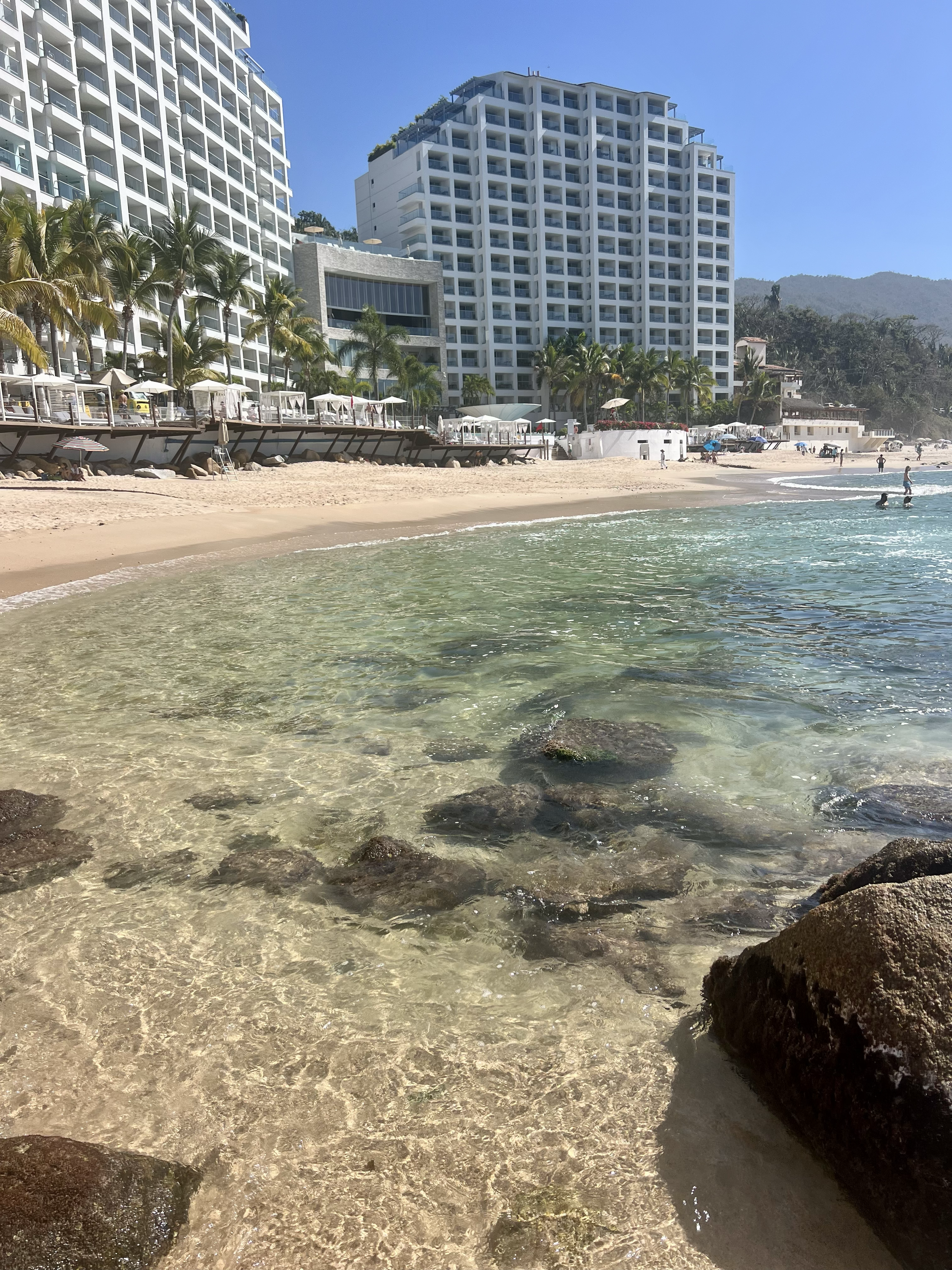A beach with clear shallow water in the foreground, and a sandy shore with people in the background. Tall modern white buildings with balconies, lush trees, and mountains are visible behind the beach on a sunny day with a blue sky.