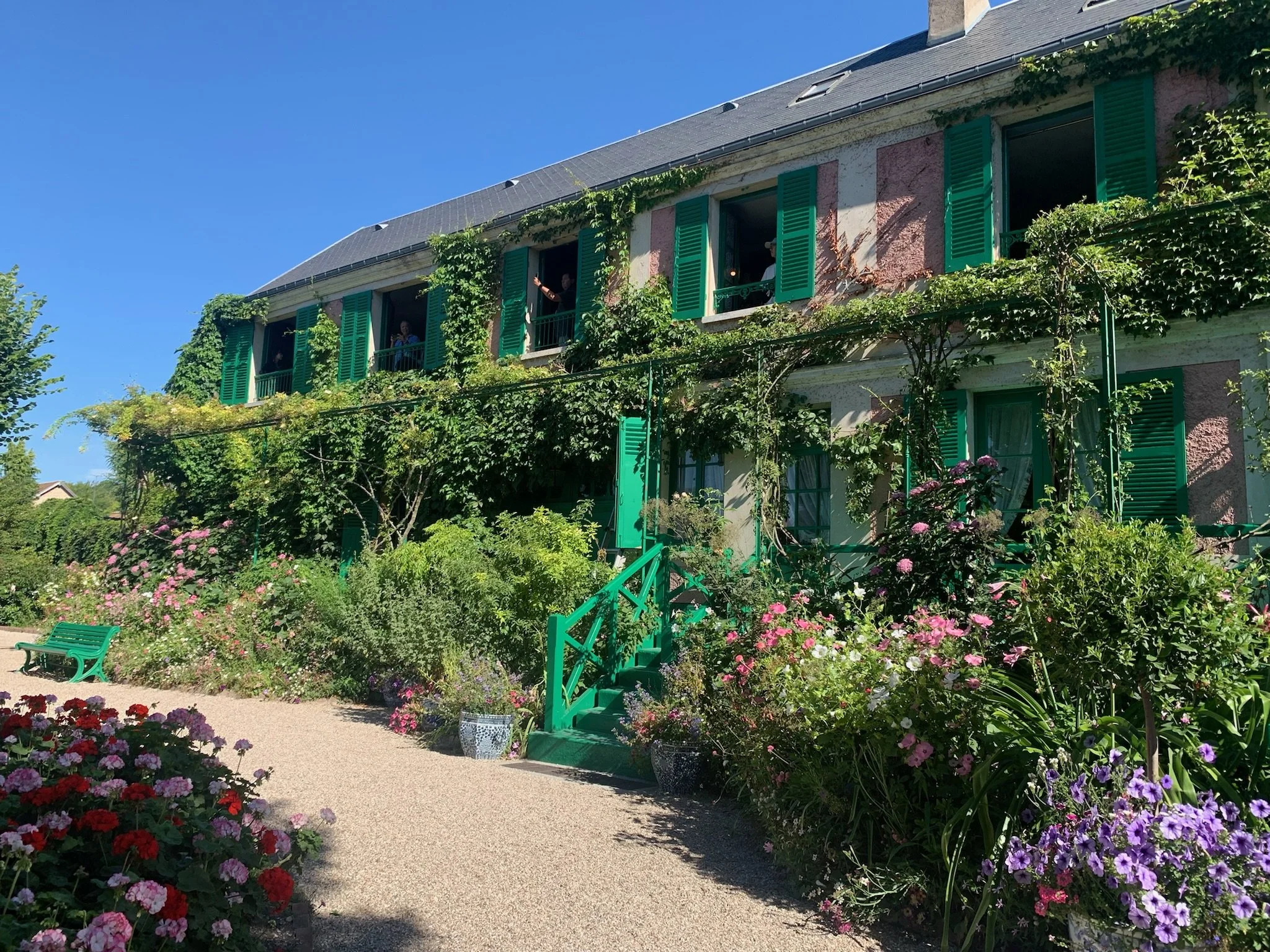 A picturesque garden with vibrant blooming flowers along a gravel path leading to a house with green shutters, vines climbing the walls, and a clear blue sky overhead.