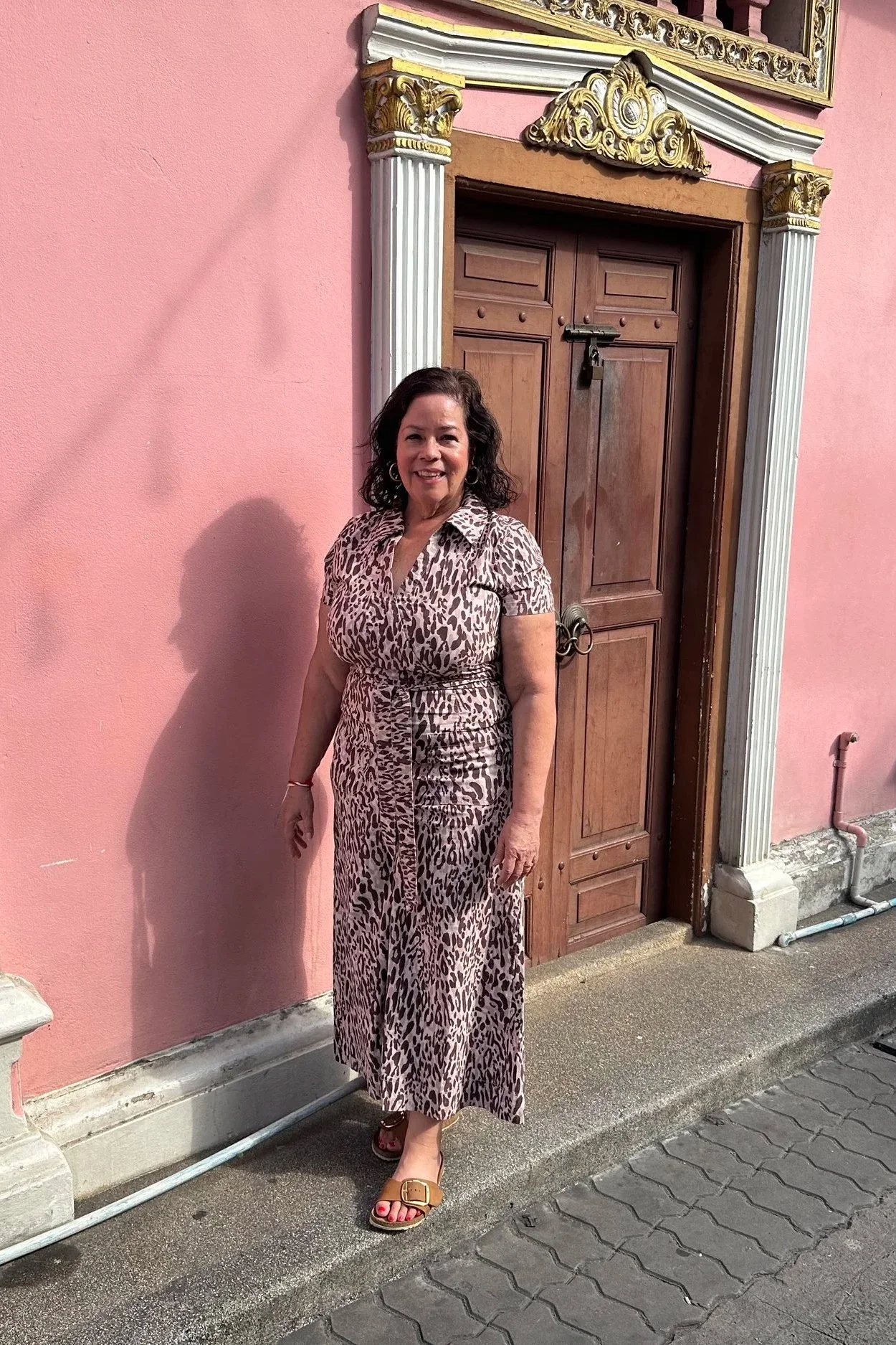 Woman standing in front of a pink wall with an ornate wooden door. She is wearing a leopard print dress and tan sandals, smiling at the camera.