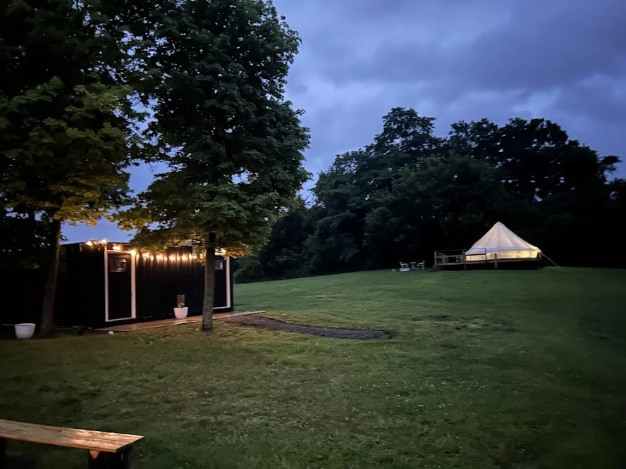 A grassy outdoor area with trees, a small black building with string lights, and a white tent on a raised platform in the background during early evening or dusk.