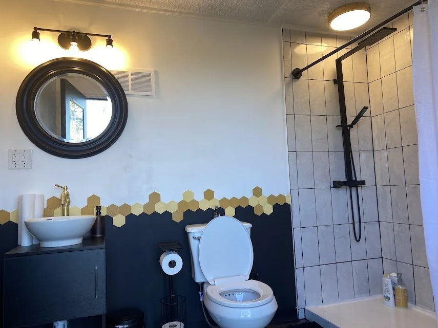 Bathroom with a black-framed round mirror, a small black vanity with a vessel sink and gold faucet, a toilet with a toilet paper holder, and a tiled walk-in shower with black fixtures and a rain shower head.