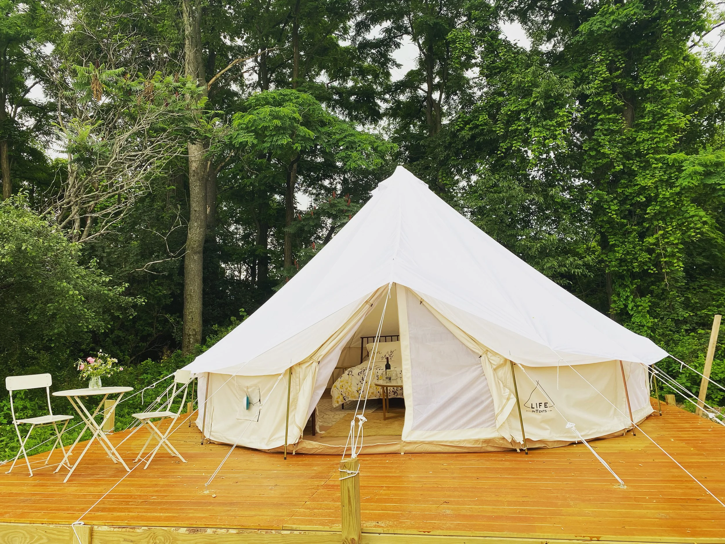 A white canvas tent with an open front, set on a wooden deck surrounded by green trees.