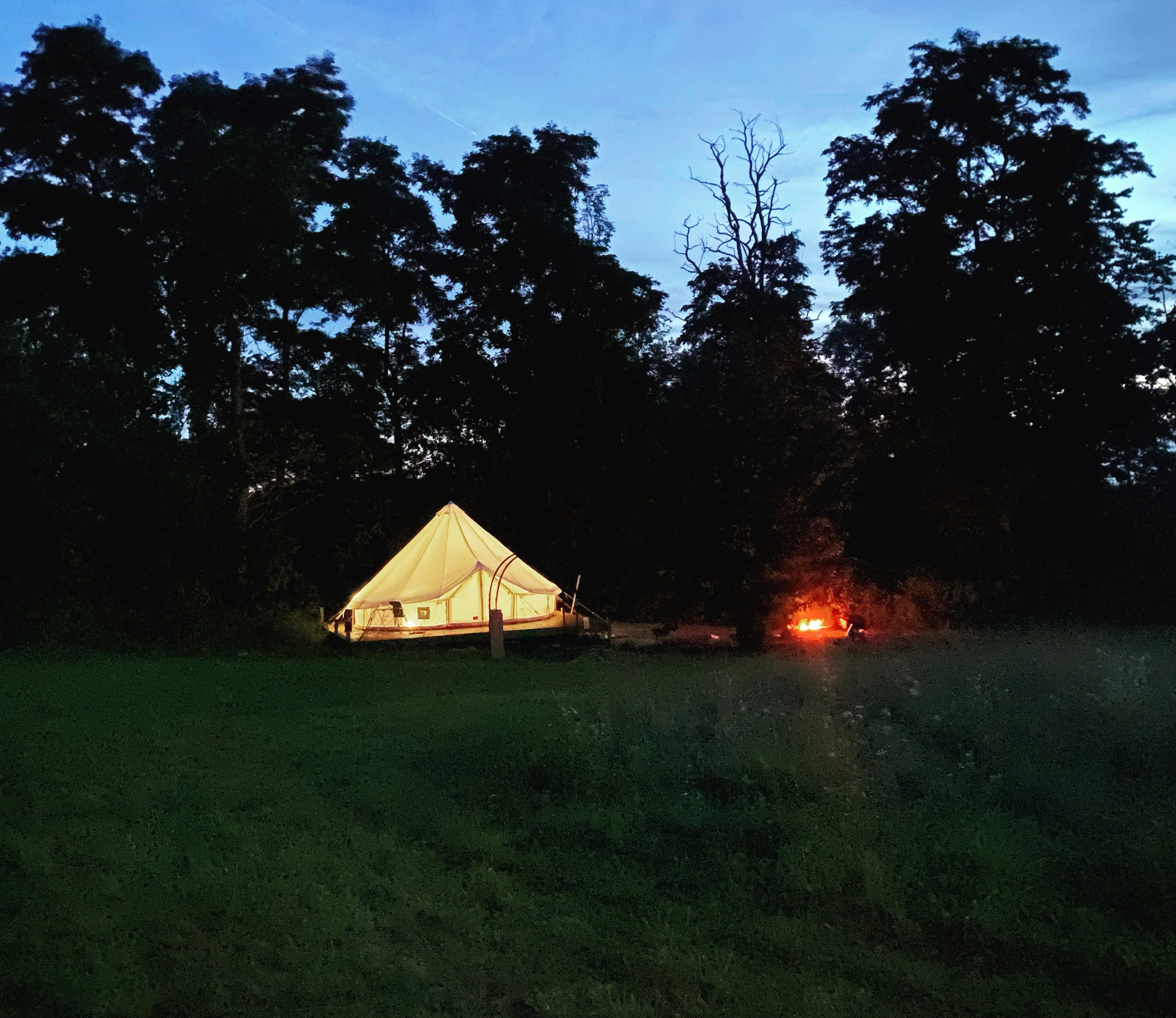 A lit tent is set up among trees during dusk, with a campfire burning nearby and a clear sky overhead.