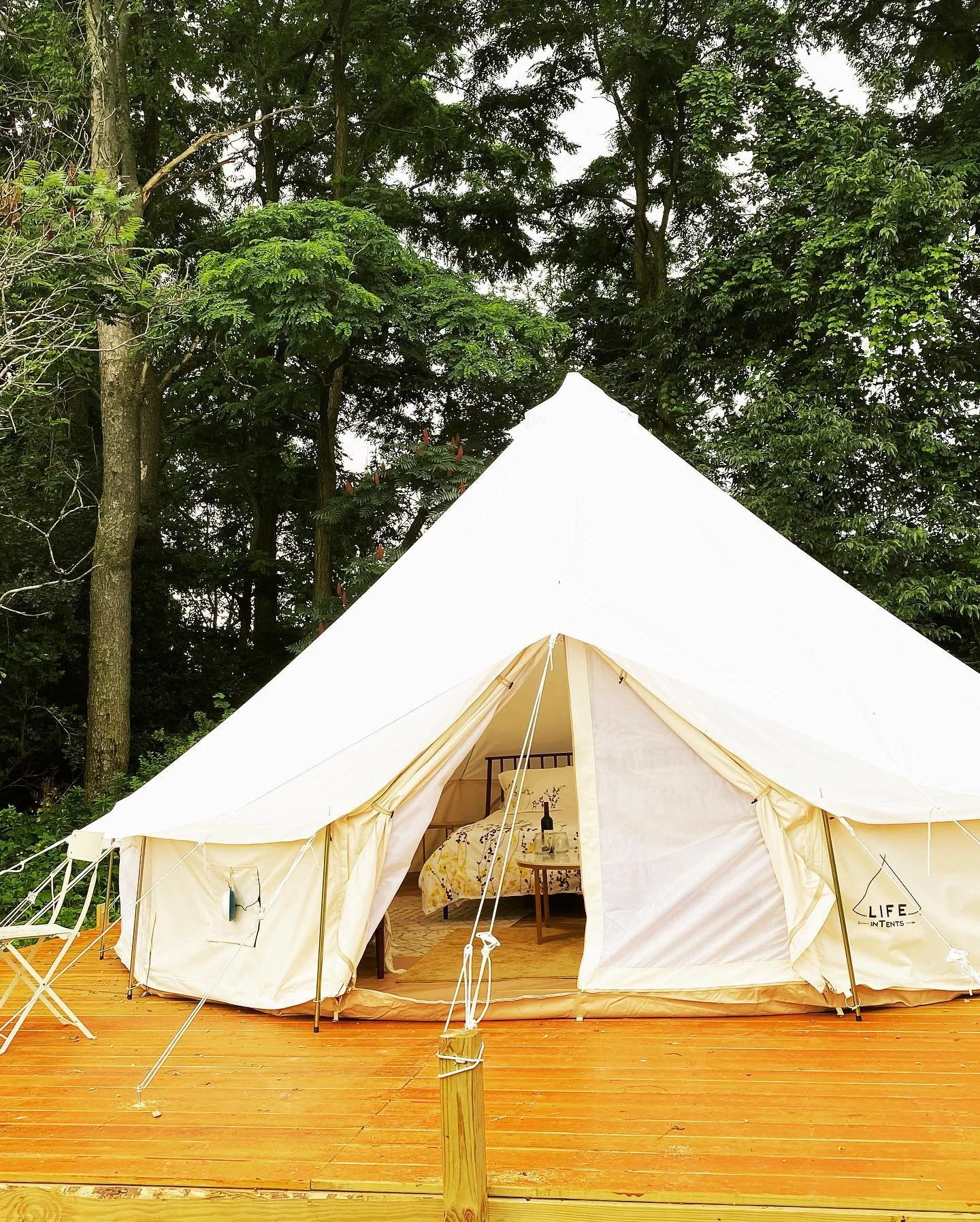 A large white canvas tent with an open door showing a bed with floral bedding inside, set on a wooden deck surrounded by trees.