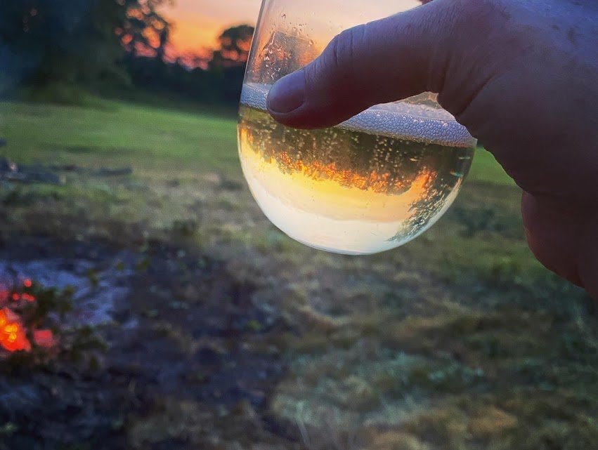 A hand holding a wine glass filled with white wine, with the sunset reflected in the glass, outdoors on a grassy area during evening.
