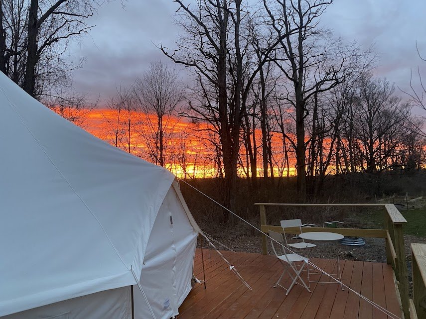 Sunset sky with vibrant orange and pink colors visible behind leafless trees, viewed from a wooden deck with a white tent, a small round table, and folding chairs.