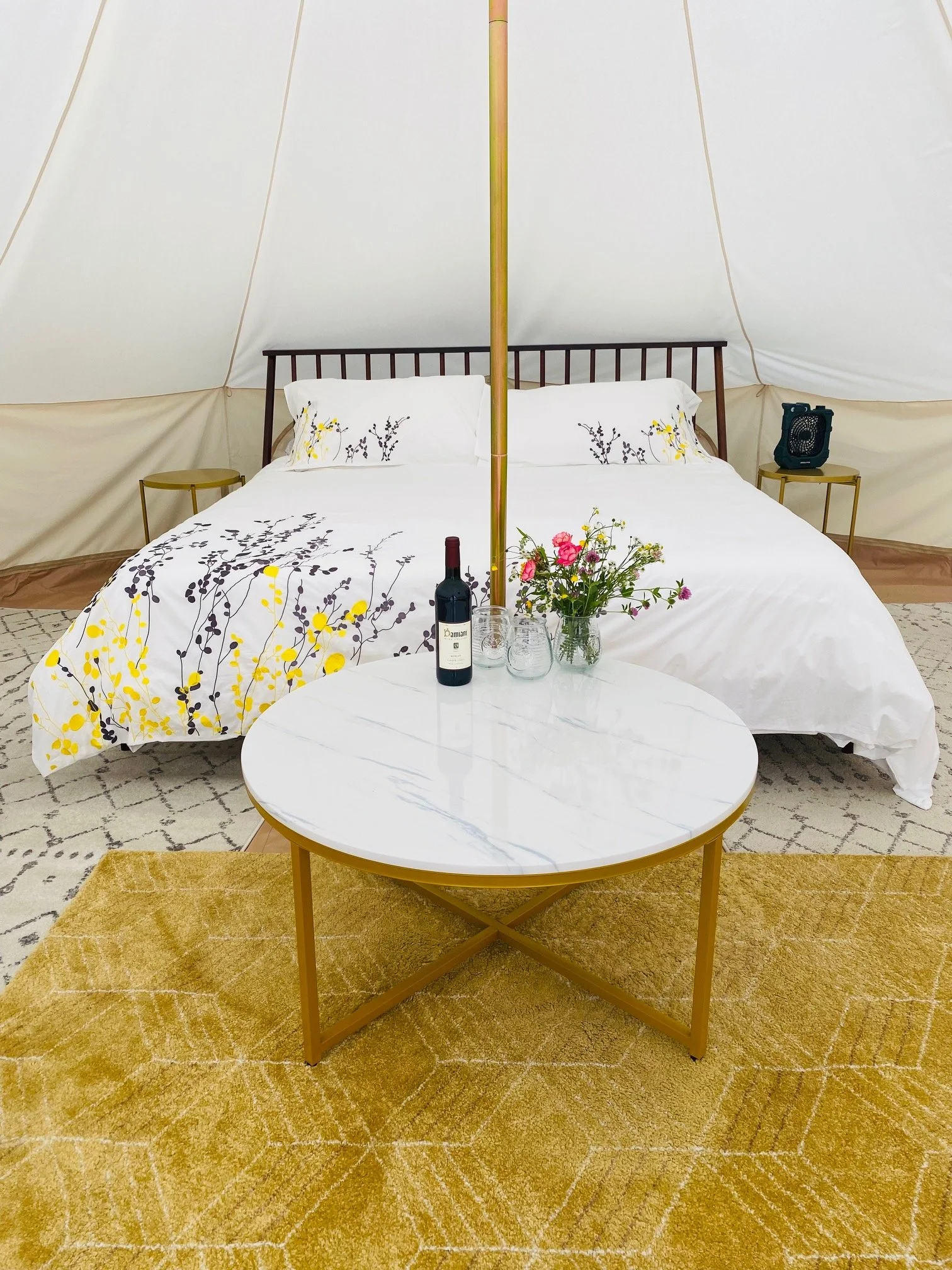 A cozy camper bed setup with a white bedspread decorated with yellow and black floral patterns, a small marble table with a bottle of wine, glasses, and a flower vase with pink and yellow flowers, in a tan and cream tent interior.
