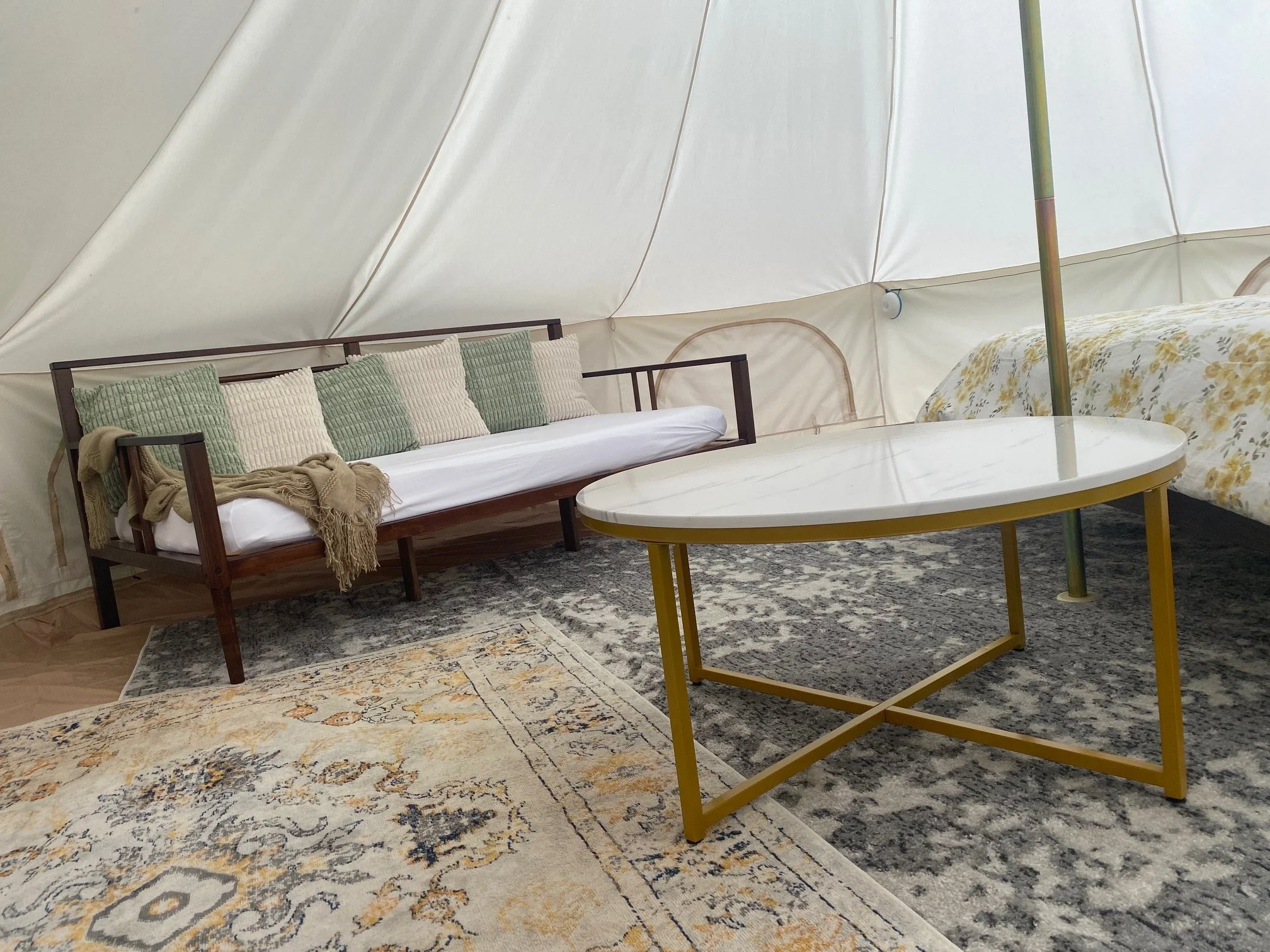 Inside a spacious tent with a wooden sofa with green and cream pillows, a round yellow table with a white top, a floral bedspread, and patterned rugs.
