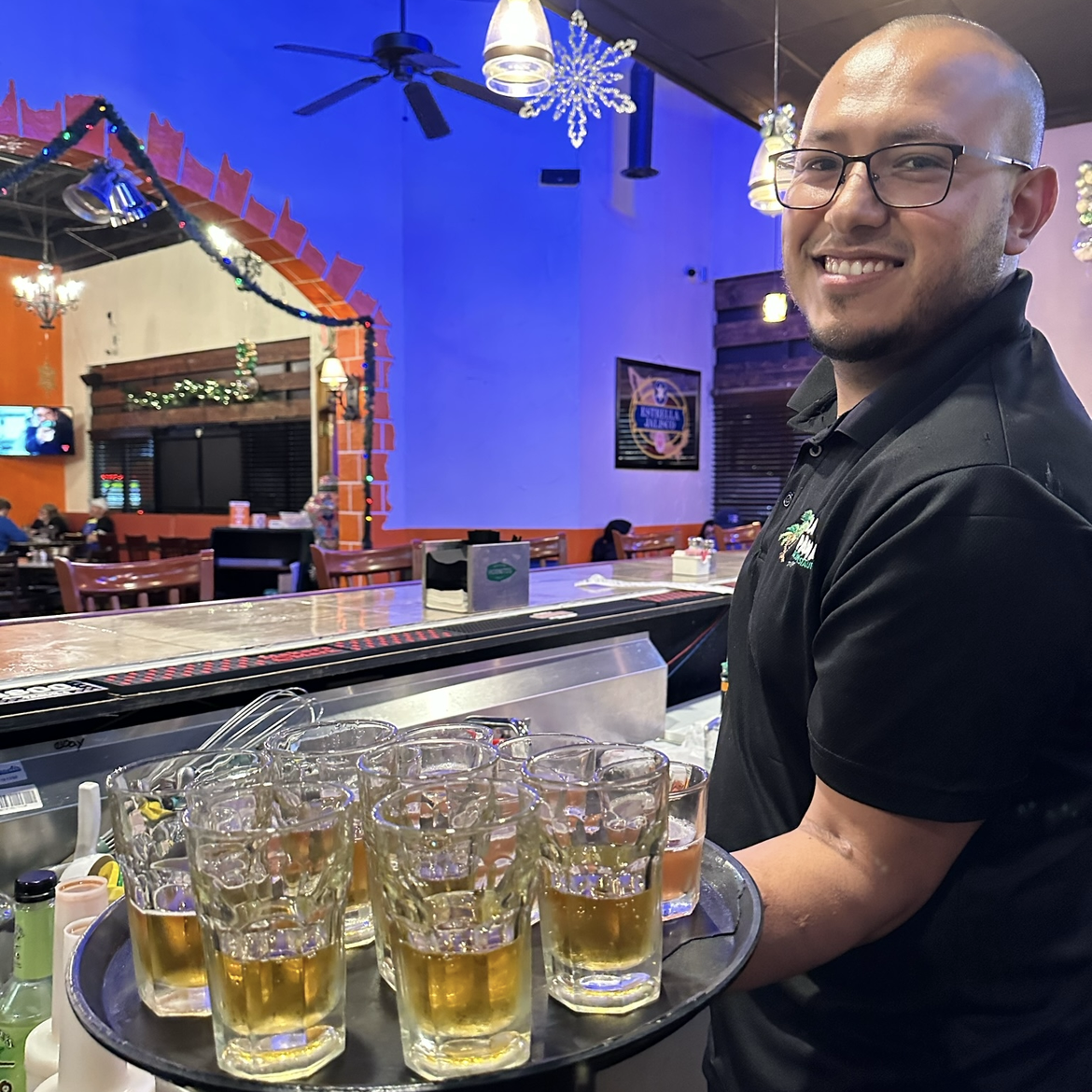 A smiling man with glasses holding a tray of glasses filled with beer inside a restaurant decorated with holiday lights and snowflake ornaments.