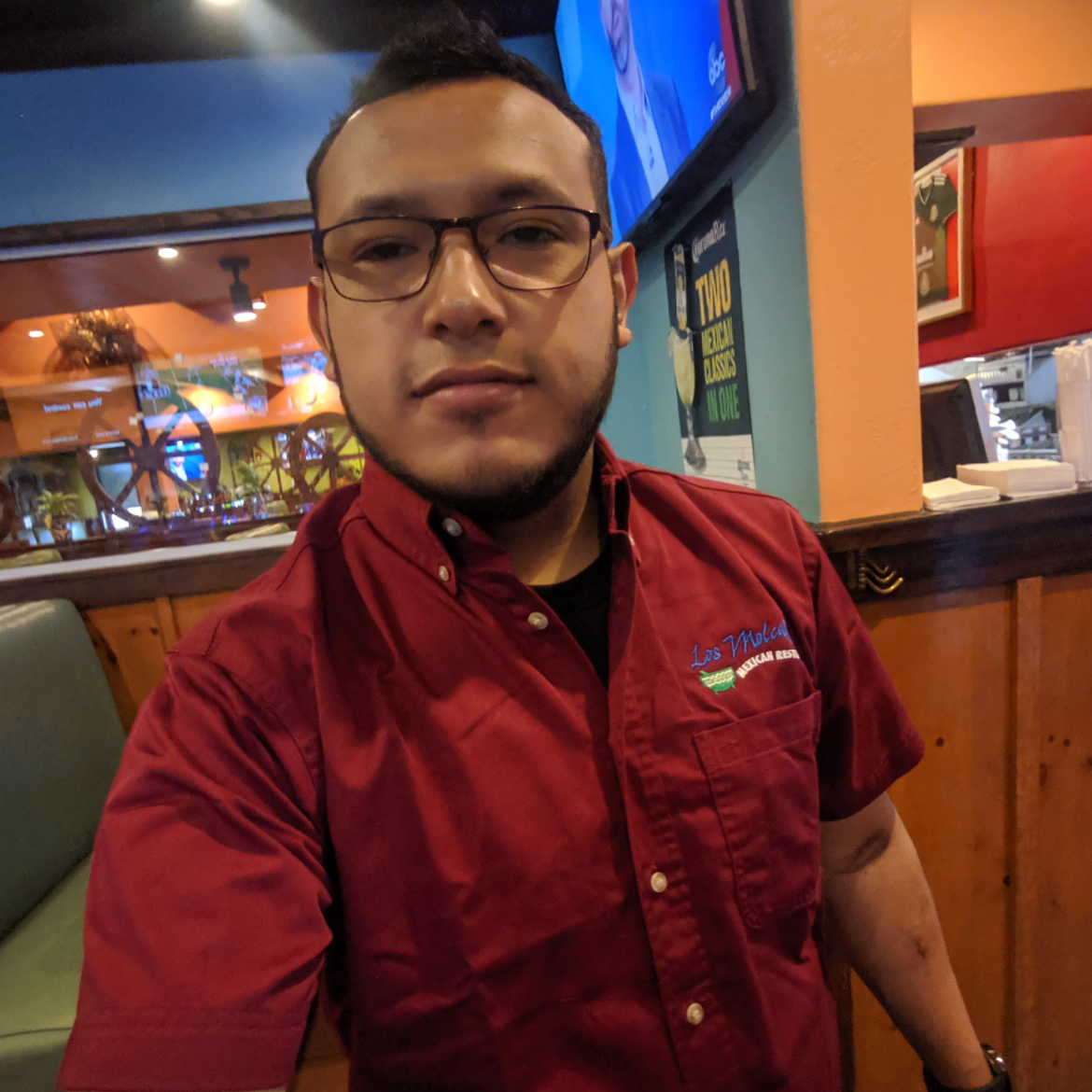 A young man wearing glasses and a red uniform shirt with the logo 'Los Molcajetes Mexican Restaurant' takes a selfie inside a restaurant.