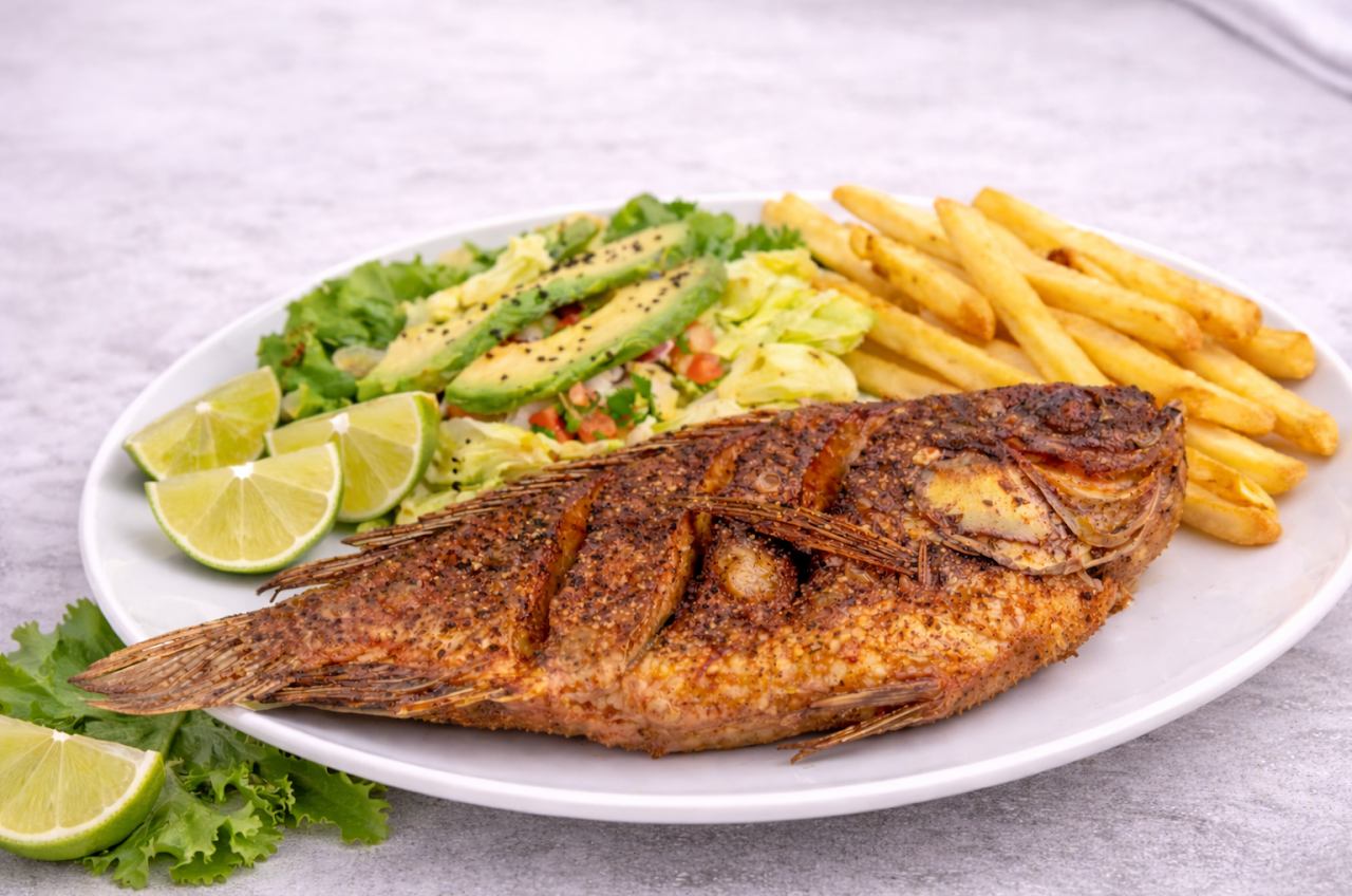 Fried whole fish with crispy skin served with French fries, avocado slices, lime wedges, and a mixed green salad on a white plate.