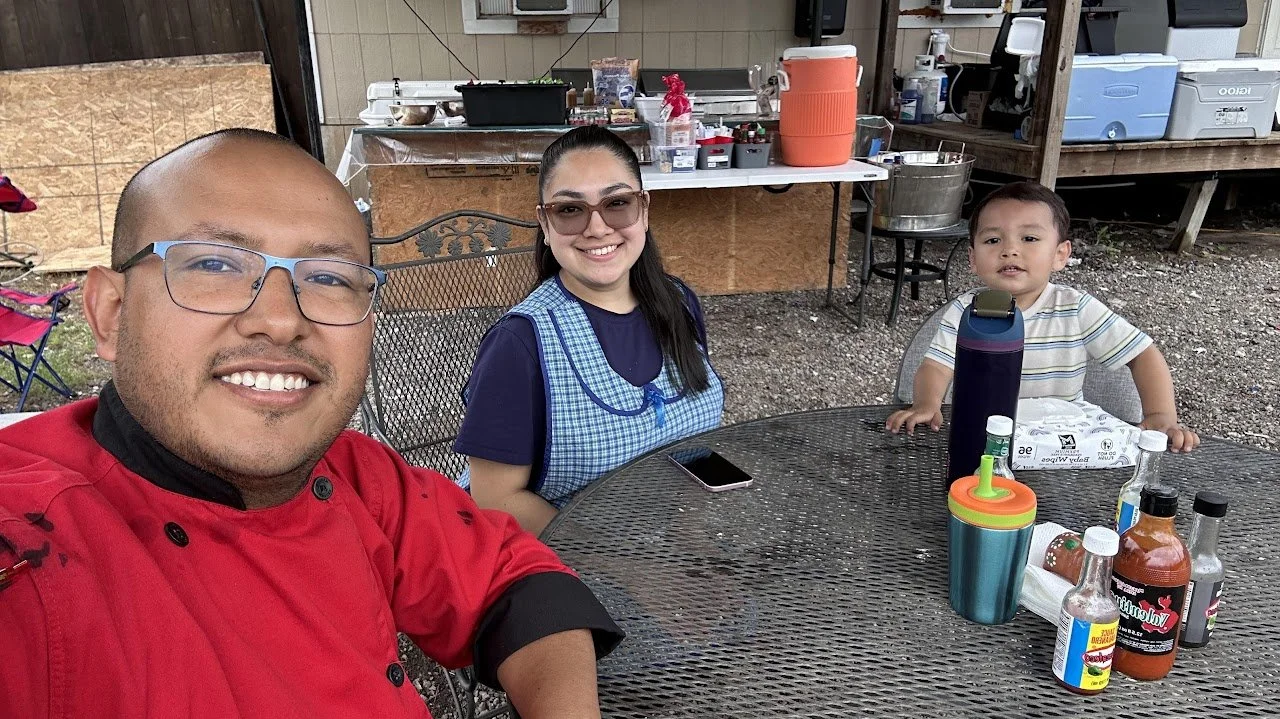 A man, woman, and young boy sitting at an outdoor metal table smiling for a selfie during a casual gathering. The man wears glasses and a red shirt, the woman has glasses and a blue apron, and the boy wears a striped shirt. On the table are drinks, condiments, and bottles, with a background of a wooden shed or deck area.