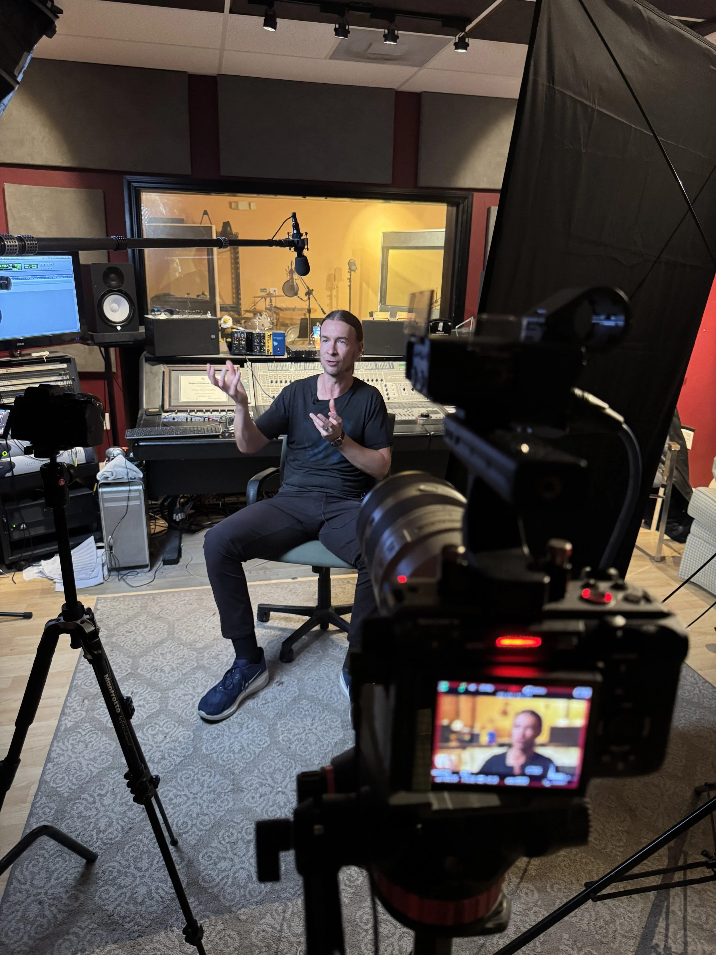 A man with long dark hair and wearing a black shirt is sitting in a recording studio, speaking and gesturing with his hands. The studio has soundproofing panels, recording equipment, and a large window looking into a live room with musical instrument