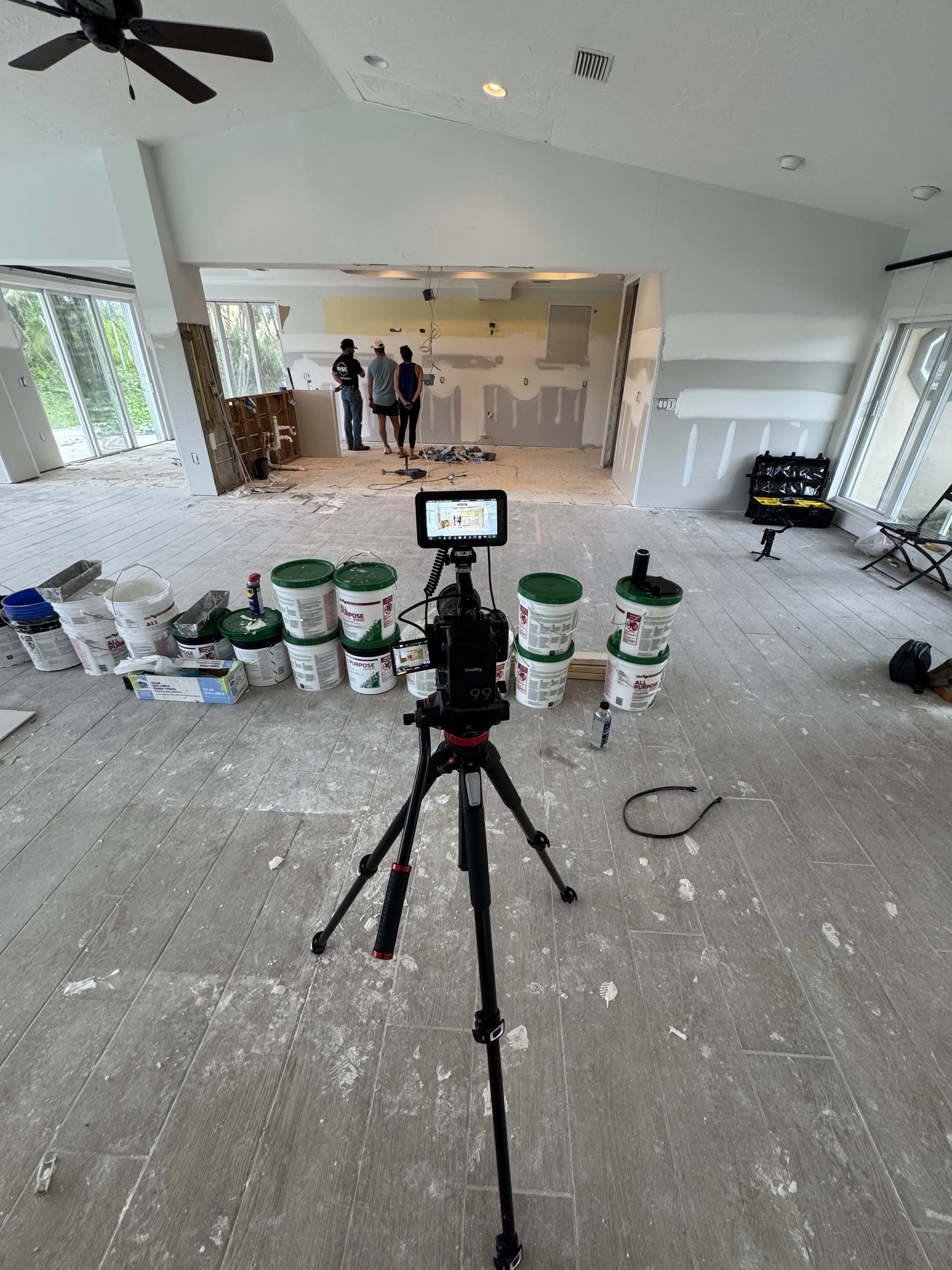 Interior of a house under renovation with construction tools, drywall, buckets, and three people discussing in the background. The photo is taken from behind a camera tripod.