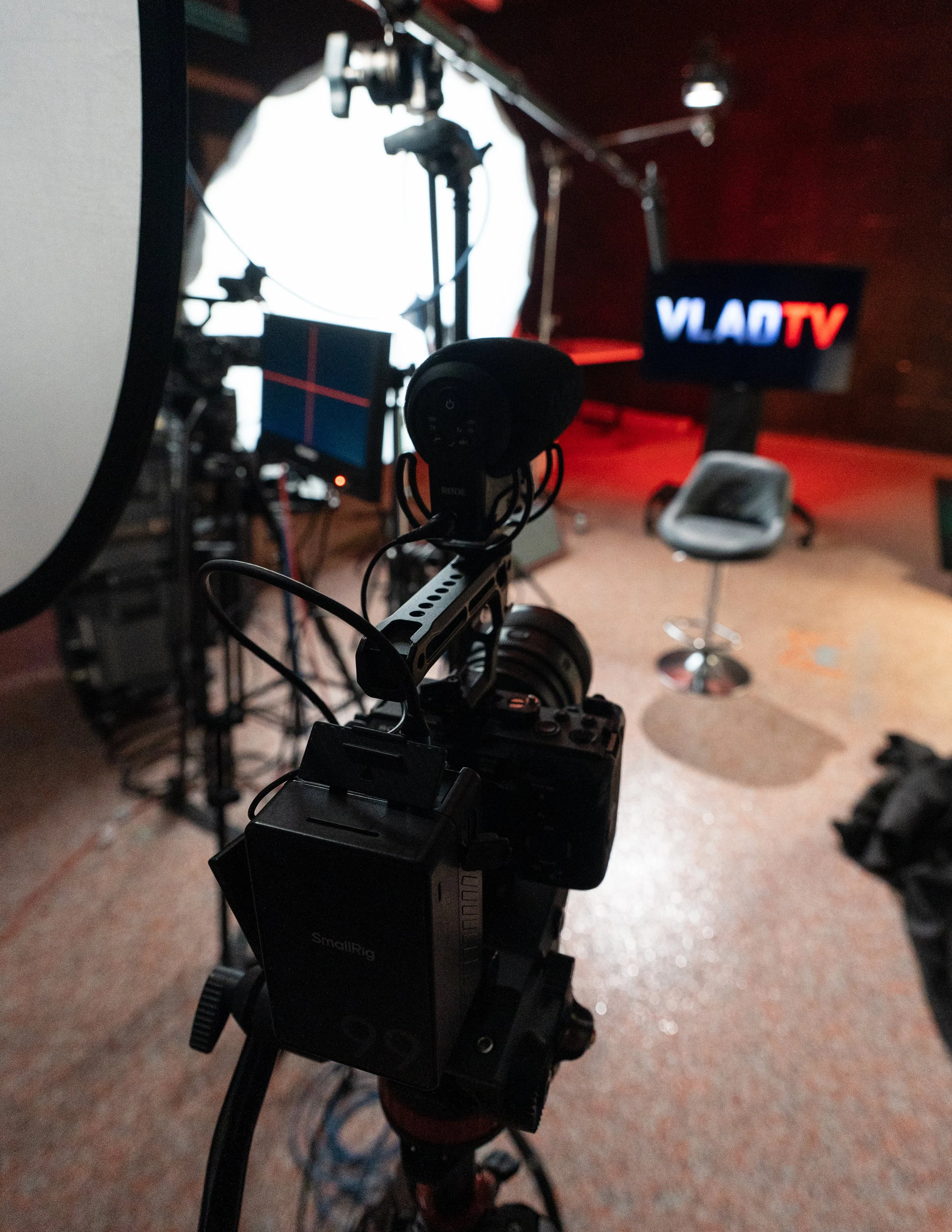 A camera on a tripod with headphones hanging from it in front of a TV screen displaying 'VLAD TV' in a studio setting with a chair nearby and various equipment in the background.
