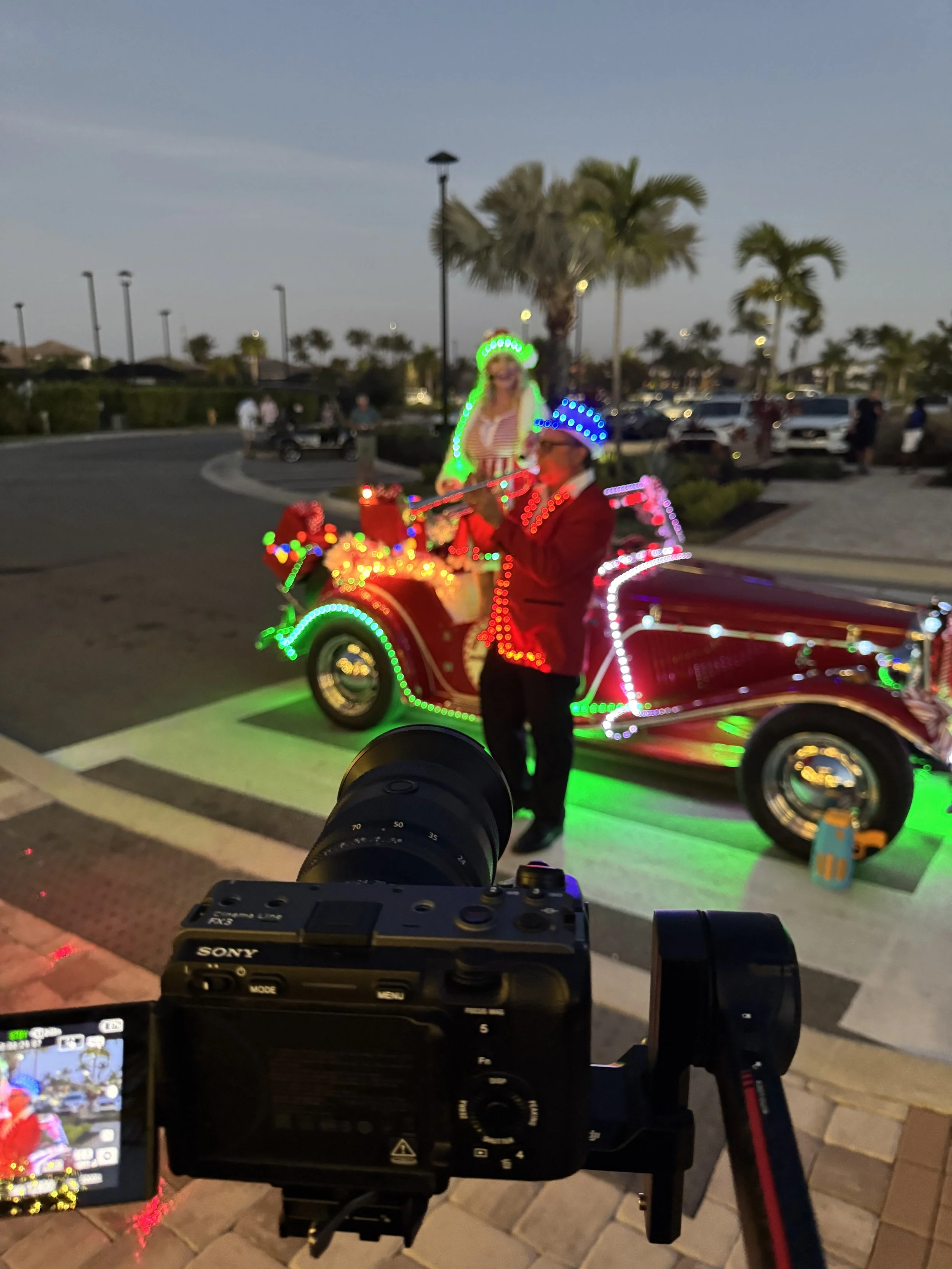 People celebrating Christmas outdoors at night, with a woman standing in front of a decorated vehicle with colorful lights, and a man enjoying the parade.