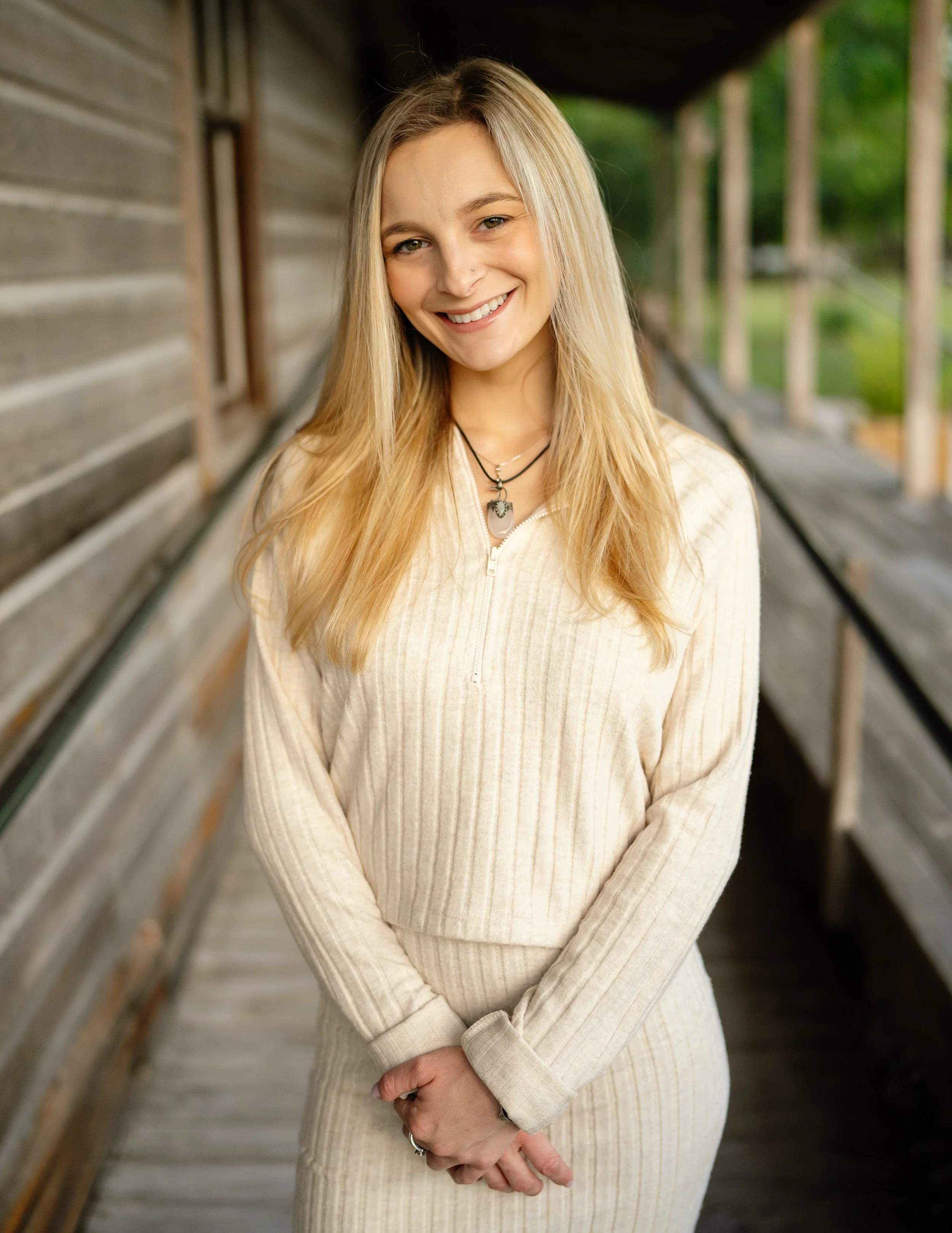A young woman with blonde hair smiling, standing outdoors on a wooden walkway, wearing a cream-colored zip-up sweater and layered necklaces.